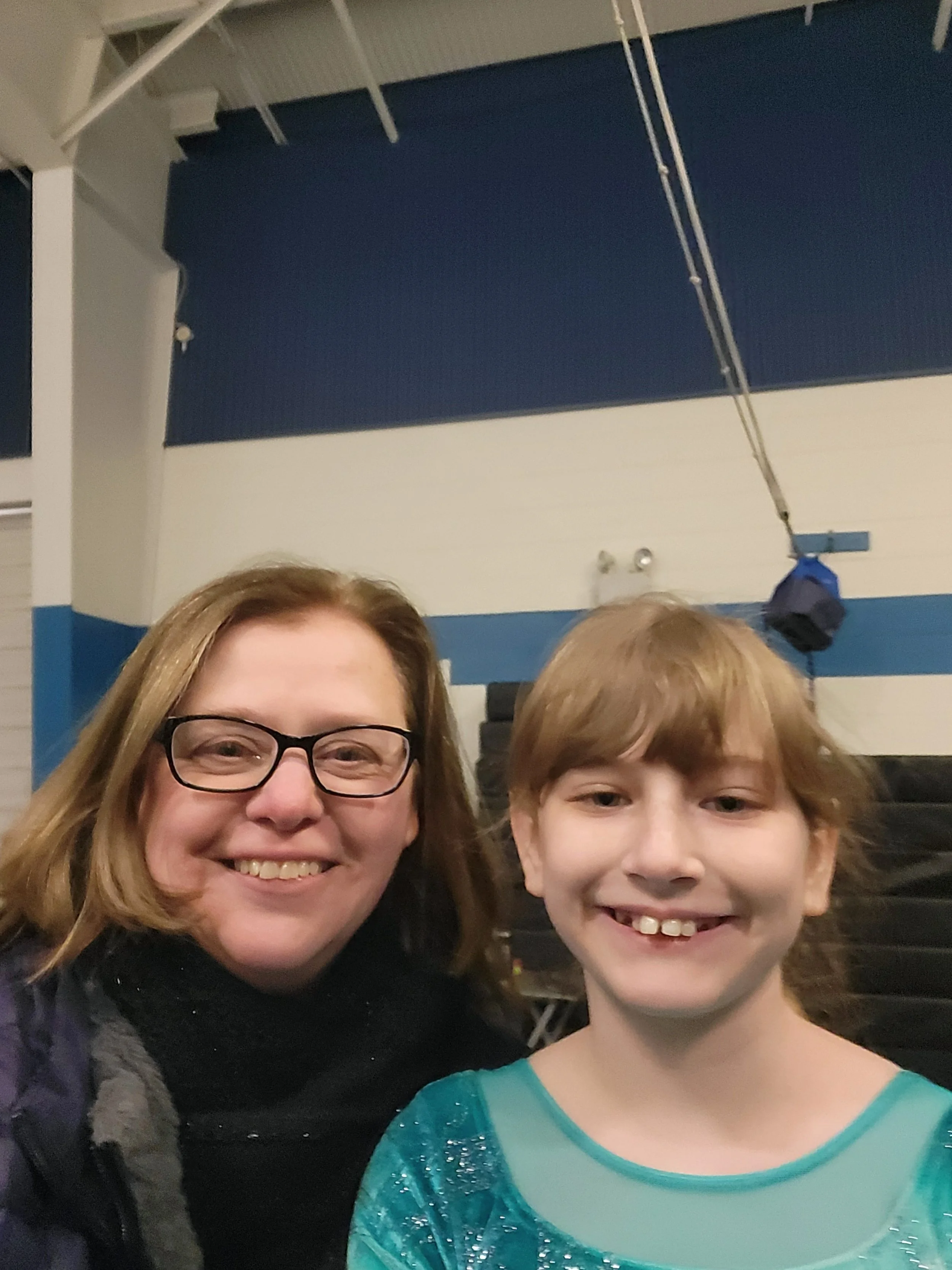 A smiling woman with glasses and a young girl with brown hair in a turquoise sparkling top are posing for a selfie in an indoor gymnasium, with blue and white walls and gym equipment in the background.