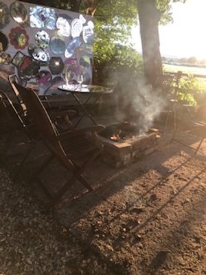 Outdoor setting with chairs around a small fire pit, artwork in the background, and trees providing shade.