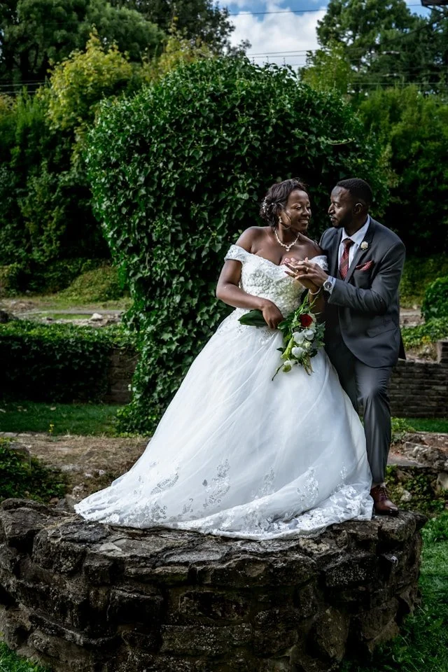 A bride and groom sharing a moment outdoors, with the bride in a white wedding gown holding a bouquet and the groom in a gray suit, standing on a stone platform in front of greenery.