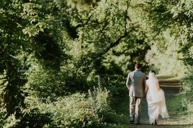 A bride and groom walking hand in hand down a shaded path surrounded by green trees.