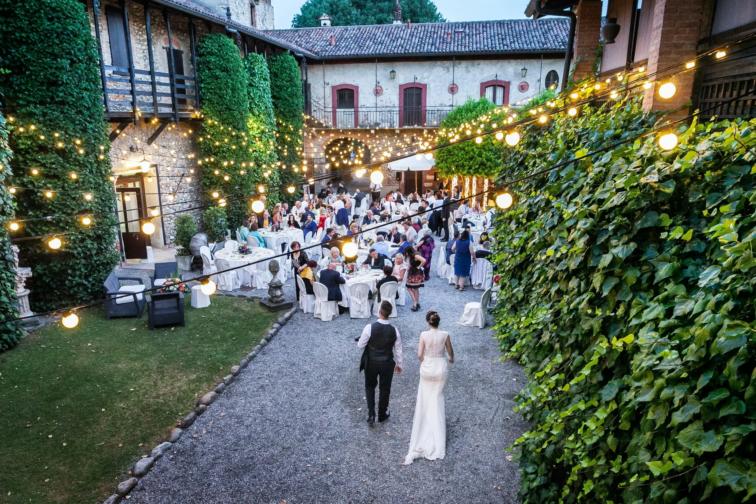 Outdoor wedding reception with string lights, round tables, and guests dining, with a bride and groom walking away from camera in evening.