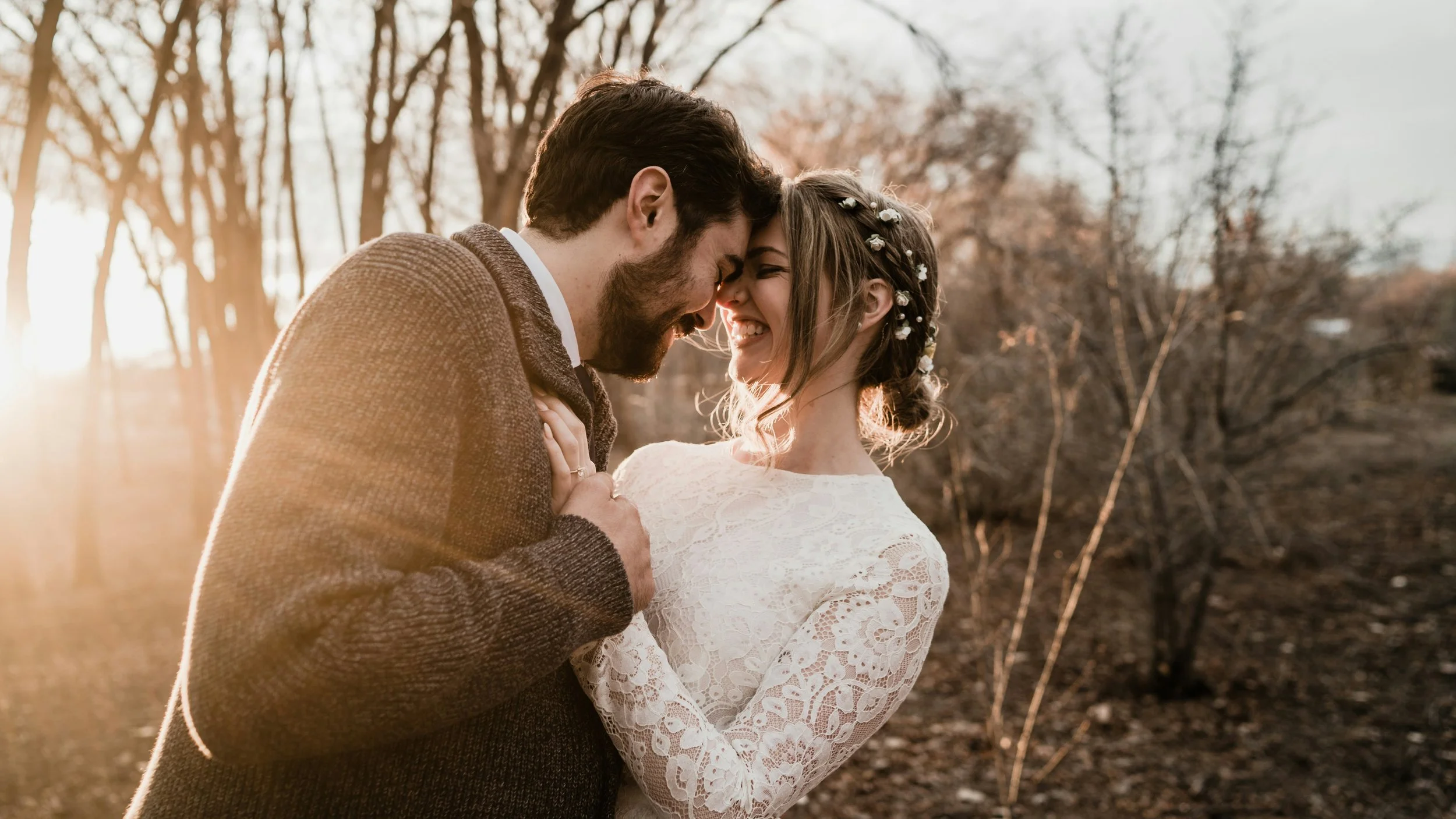 A smiling couple touching foreheads and holding hands, standing outdoors during sunset, with bare trees in the background.
