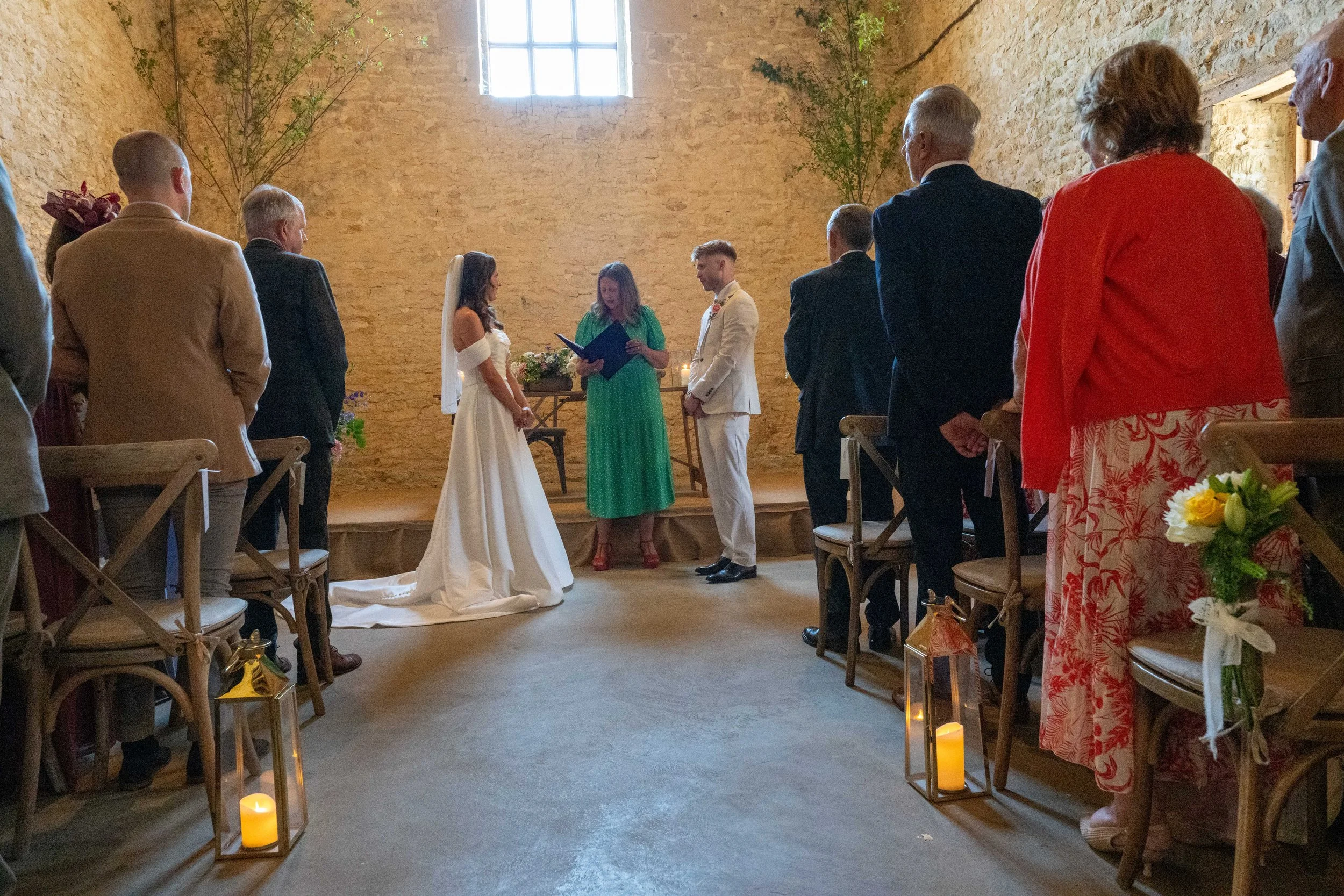 A wedding ceremony with the bride and groom standing at the altar, surrounded by guests in a rustic indoor setting with exposed brick walls, trees, and candles.
