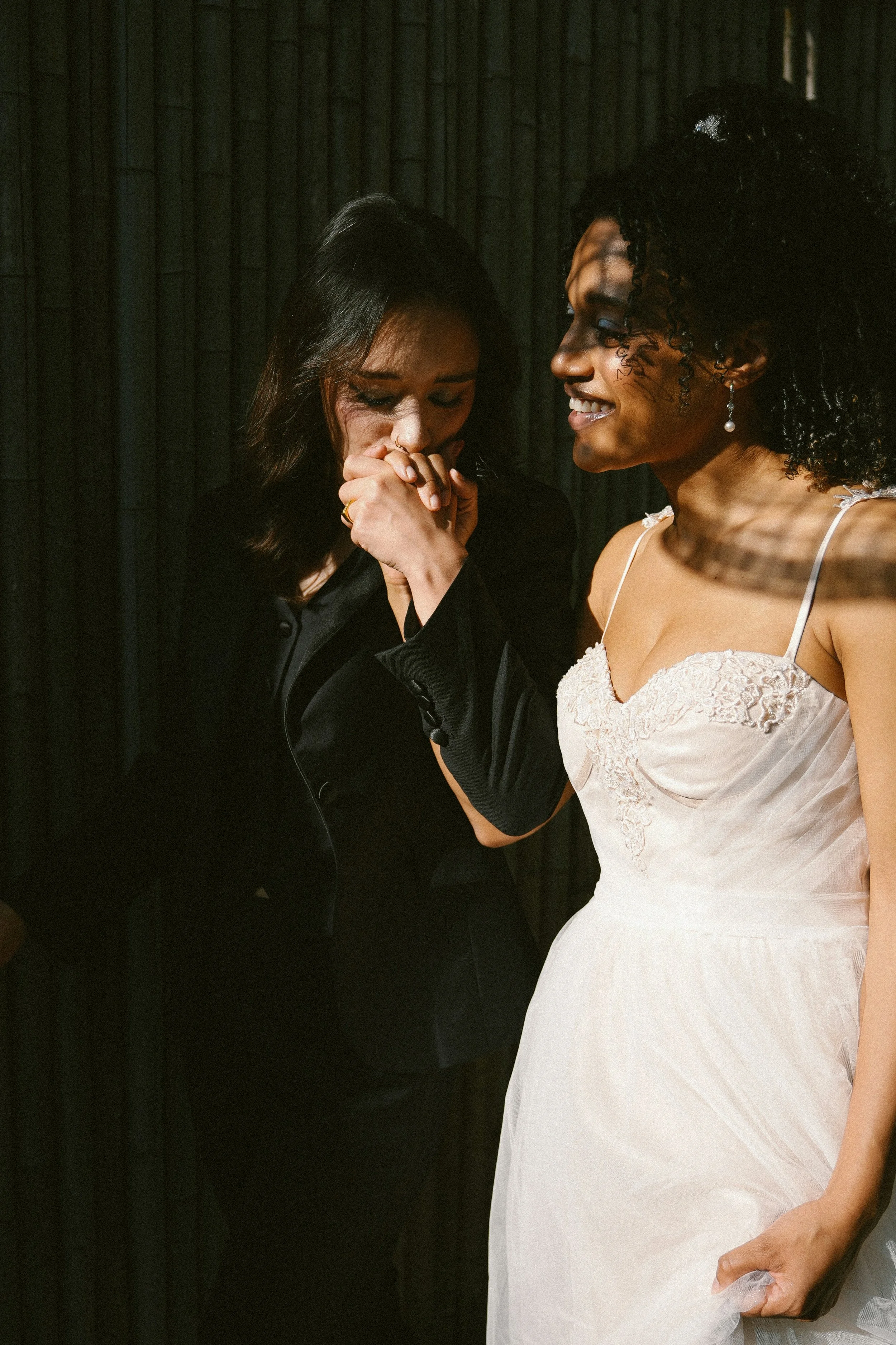 Two women share an emotional moment, with one woman dressed in a white dress and the other in black. The woman in white is smiling, while the woman in black looks touched, covering her face with her hands.