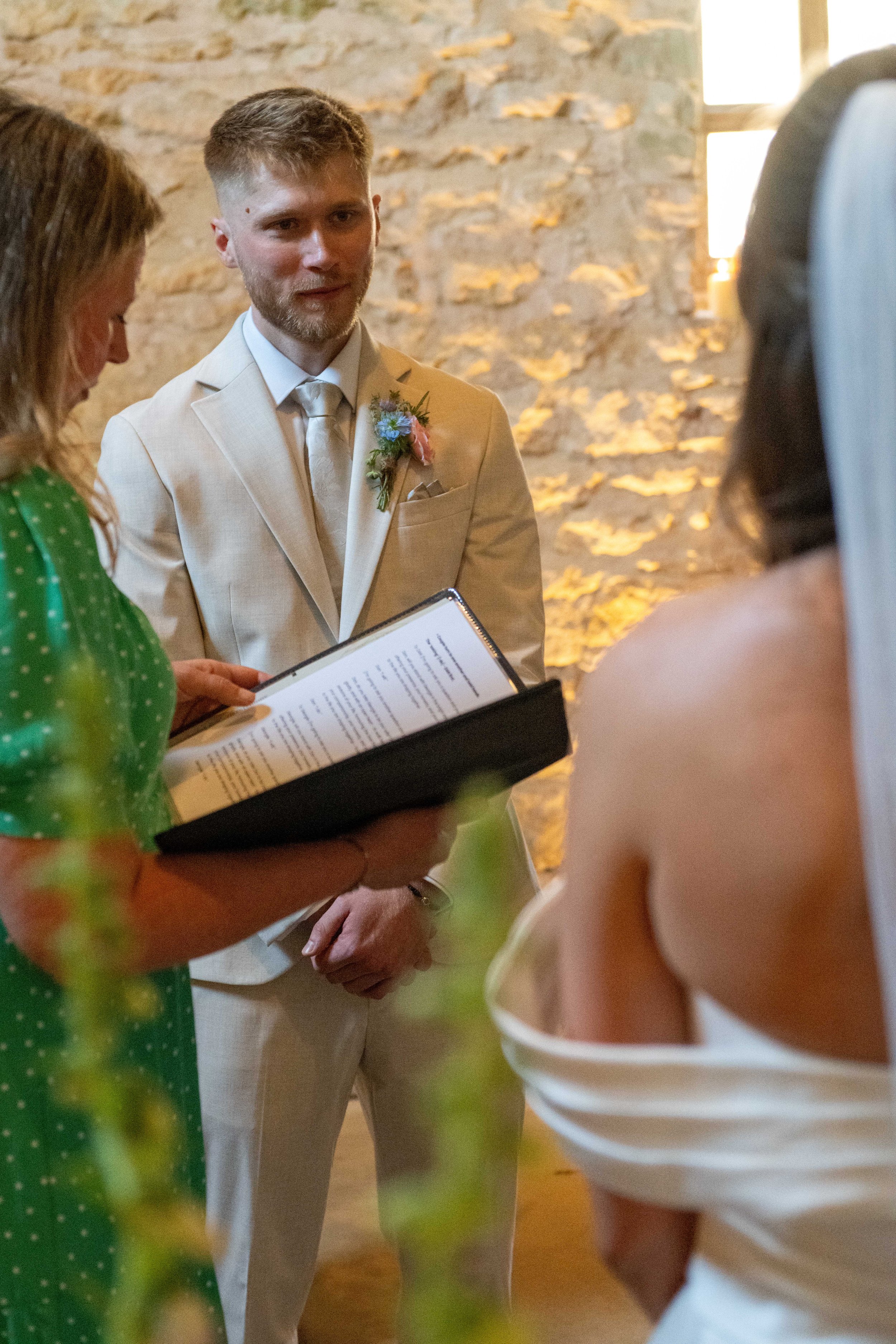 A wedding ceremony with a groom in a beige suit and a white tie, a woman in a green polka dot dress, and other people, in a rustic indoor setting with a stone wall.