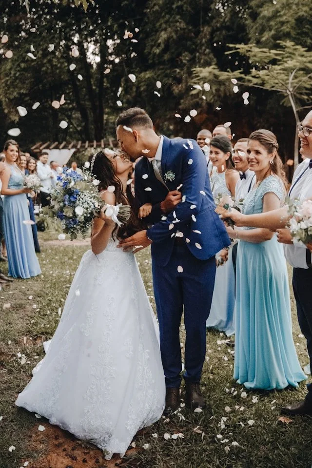 A bride and groom kissing at their outdoor wedding as guests watch and celebrate, tossing flower petals in the air.
