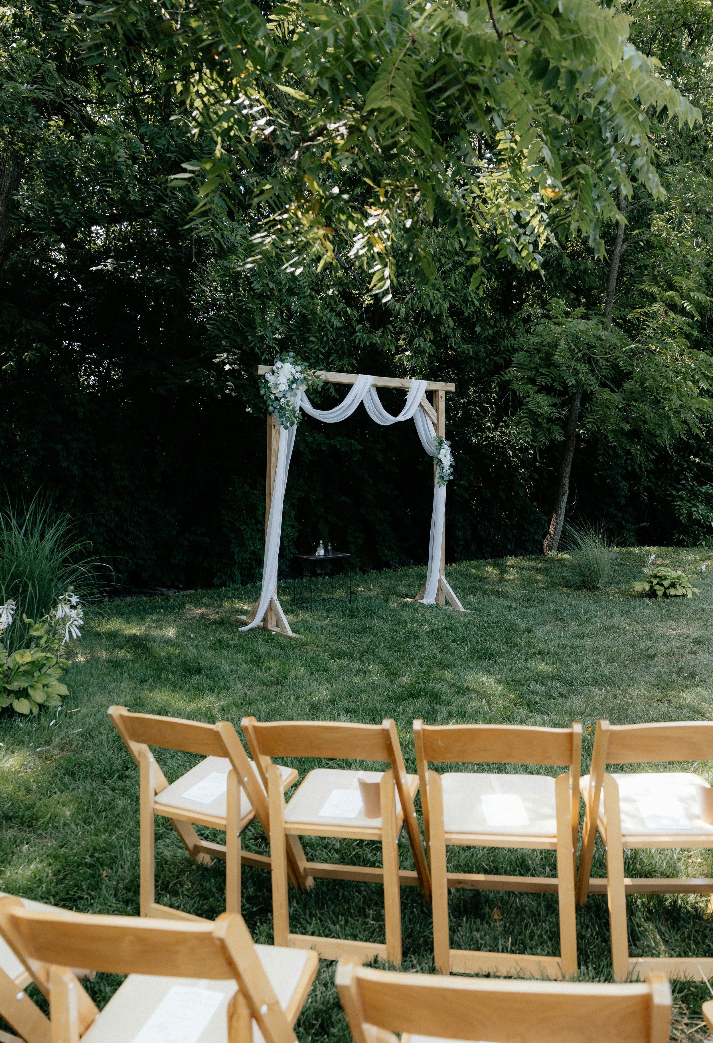 Outdoor wedding ceremony setup with wooden chairs arranged in front of a decorated wooden arch on green grass, surrounded by trees.
