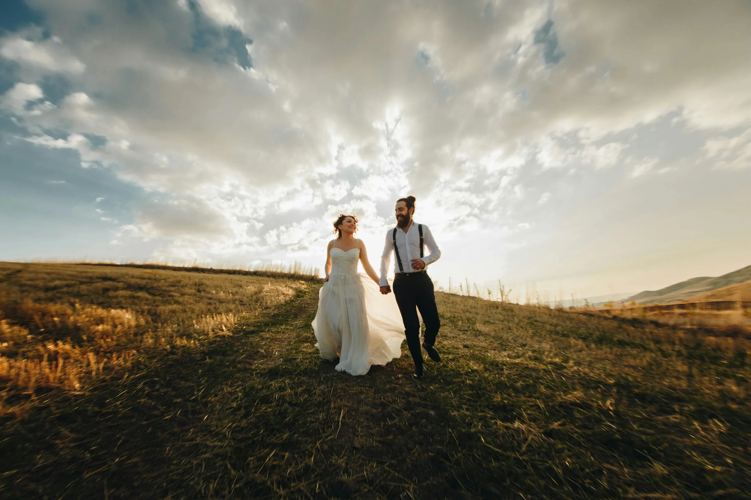 A newlywed couple walking hand in hand in a field during sunset, with a dramatic sky filled with clouds overhead.
