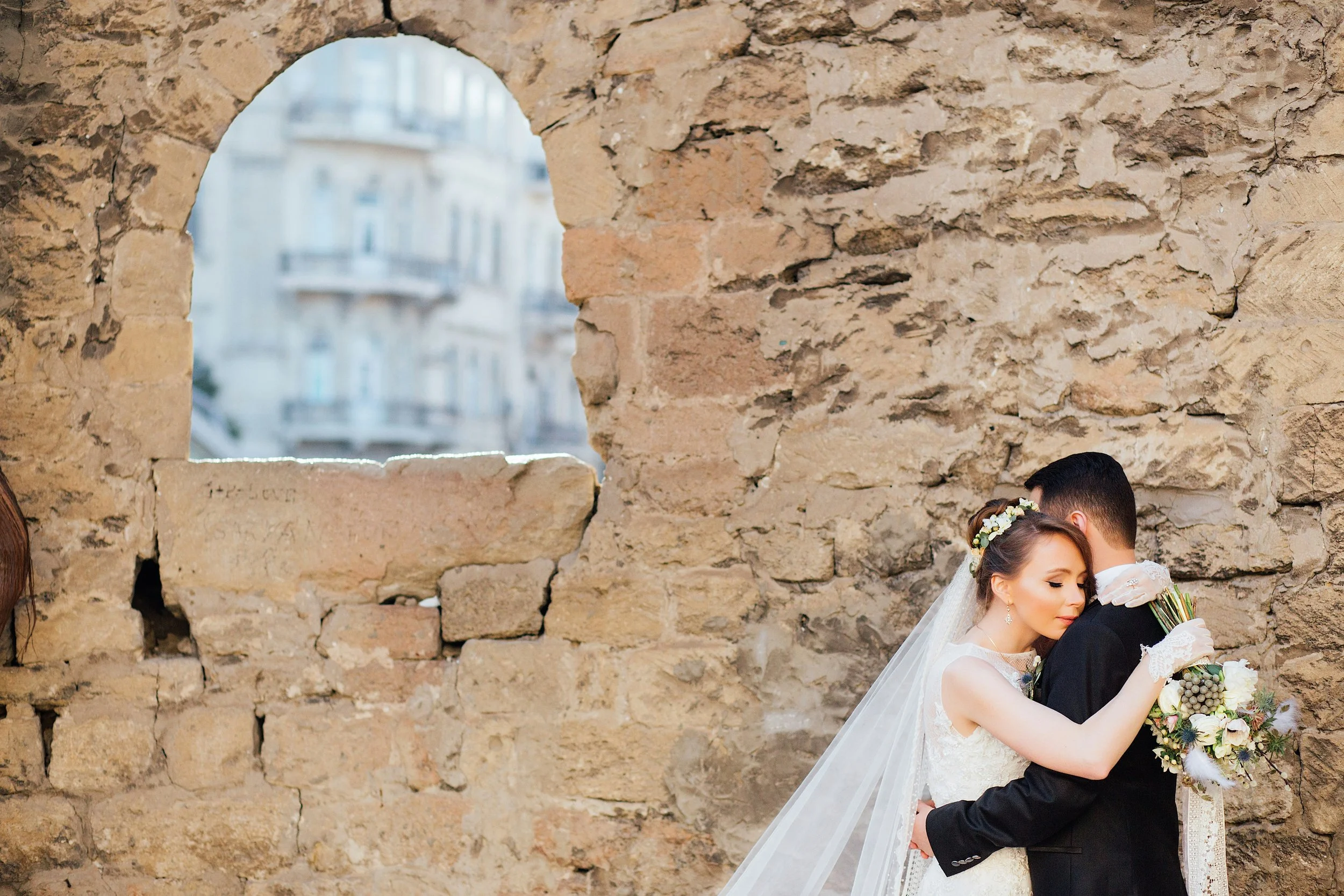 A bride and groom embracing against an old stone wall, with the bride wearing a white wedding gown and veil, and the groom in a black suit, holding a bouquet of flowers.