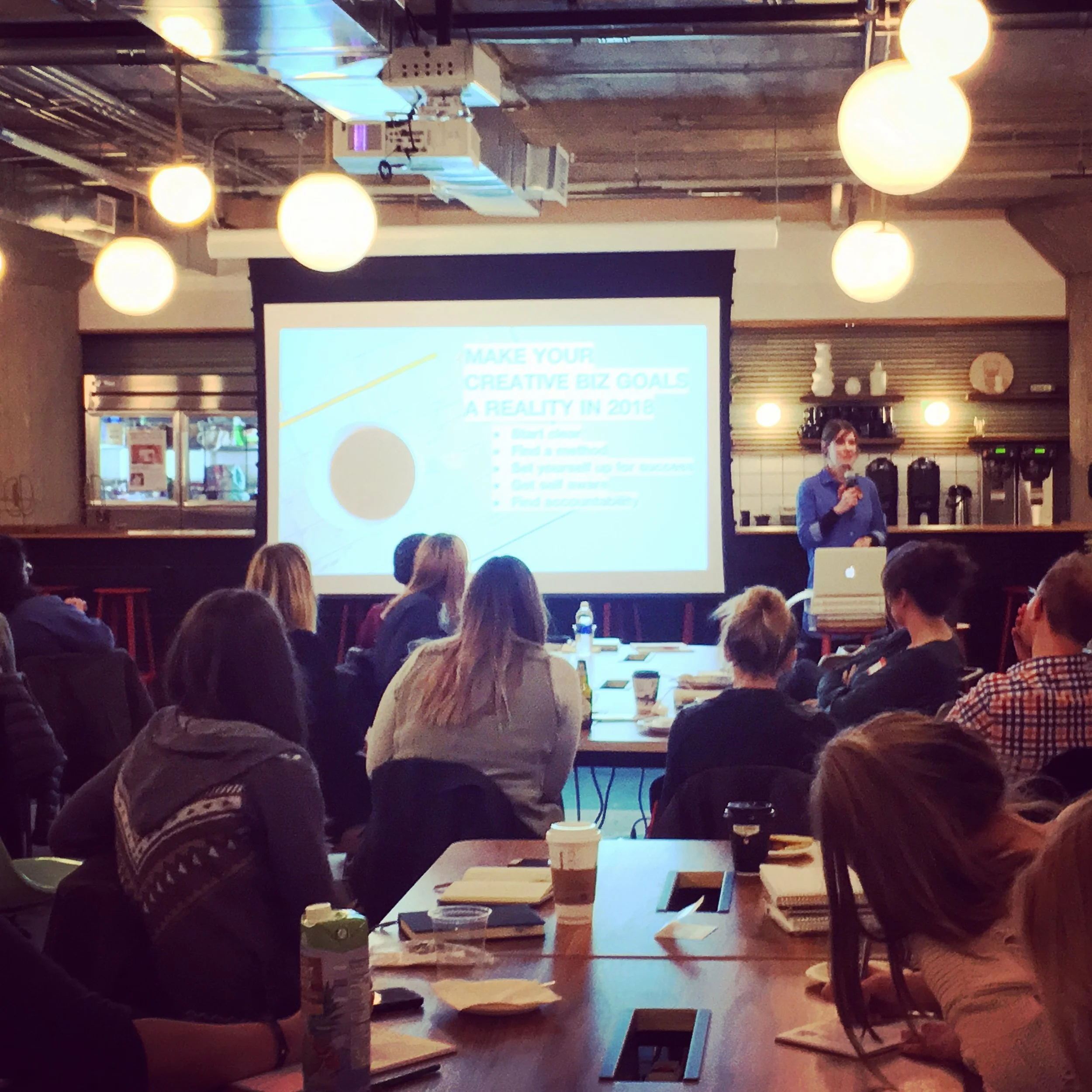 A woman giving a presentation to a group of people in a restaurant or cafe with a large screen behind her displaying a PowerPoint slide titled 'MAKE YOUR CREATIVE BIZ GOALS A REALITY IN 2018.' The audience is seated at tables with notebooks, cups, and water bottles.