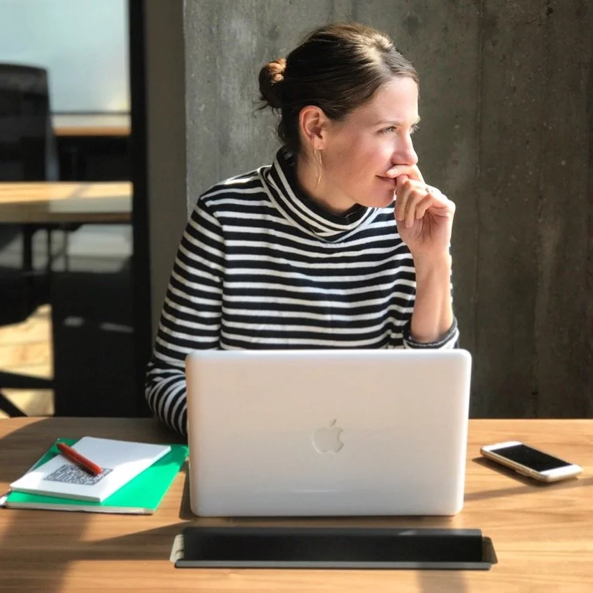 A woman with dark hair tied back, wearing a black and white striped turtleneck, sits at a wooden table with a silver MacBook, a green notebook, a red pen, and a smartphone in an office or cafe setting with a concrete wall in the background.