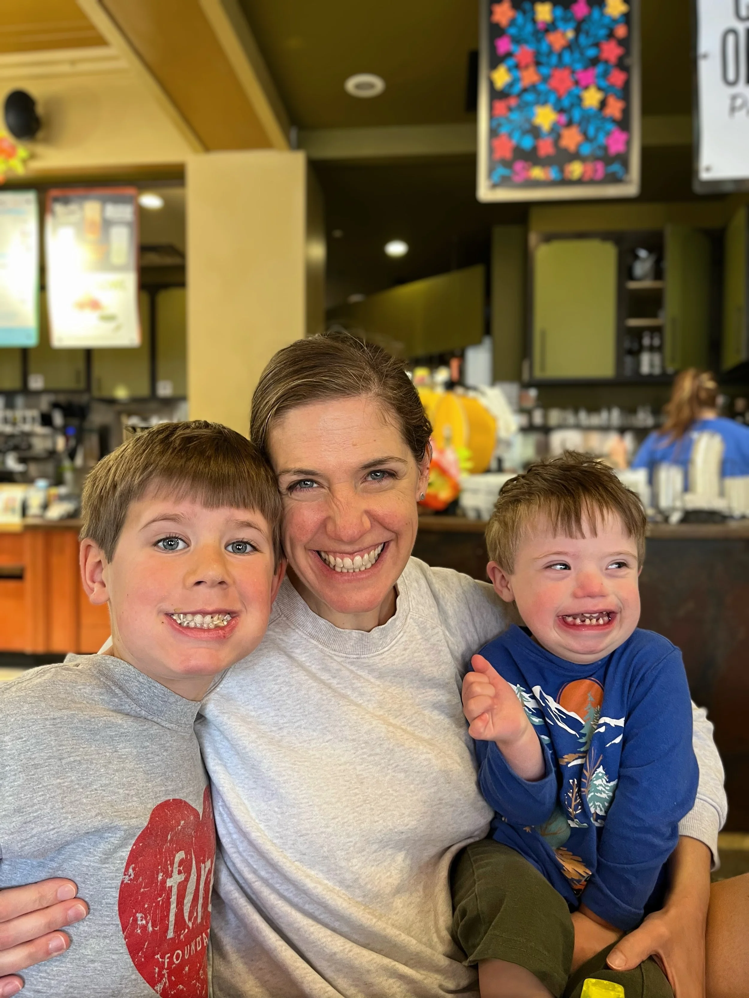 A woman smiling with two young boys, both with missing front teeth, in a cafe or restaurant setting.