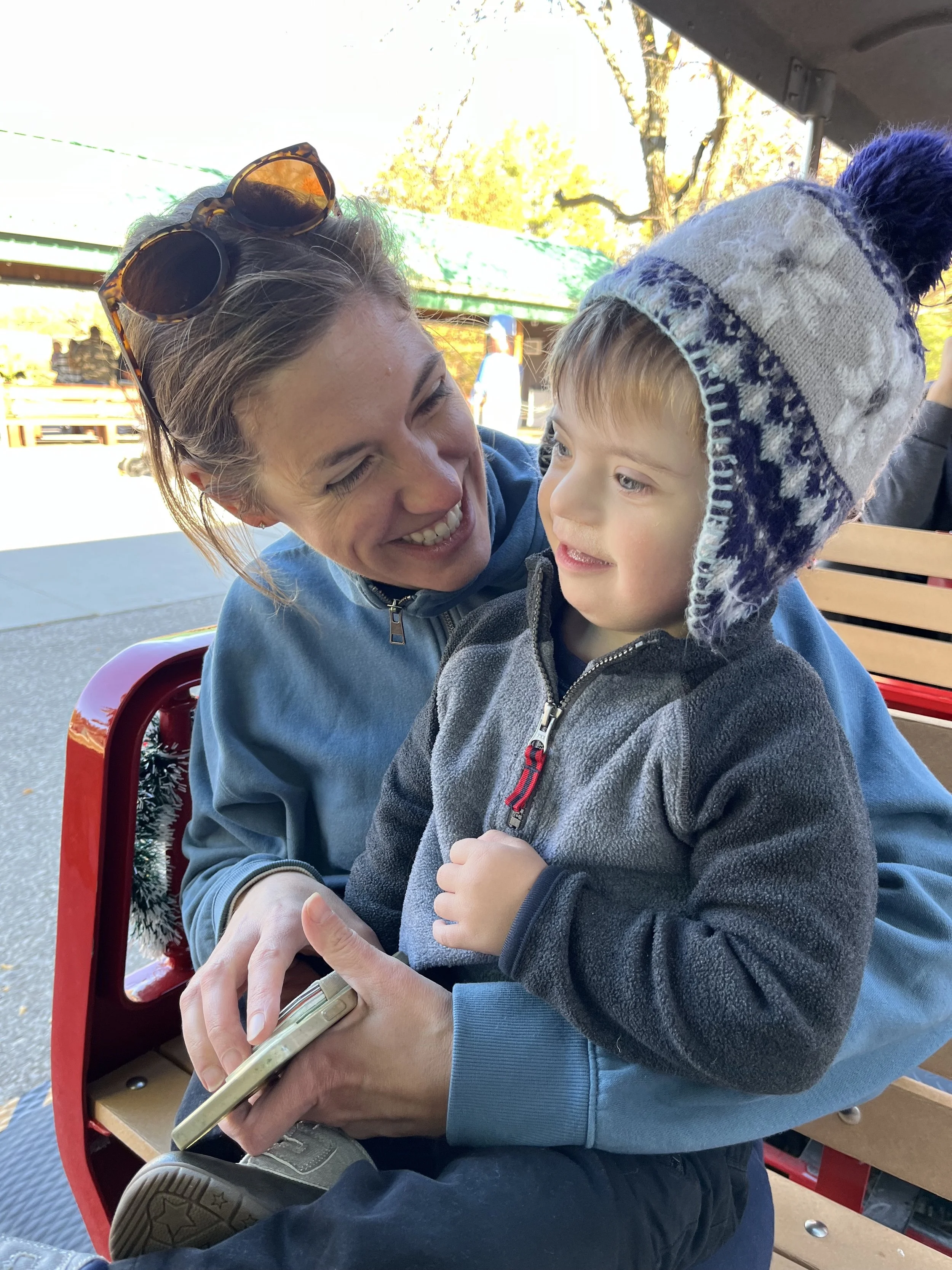 A woman and a young boy sitting together outdoors, smiling at each other. The woman is holding a smartphone and wearing sunglasses on her head, while the boy is wearing a gray and blue knit hat and a gray jacket.