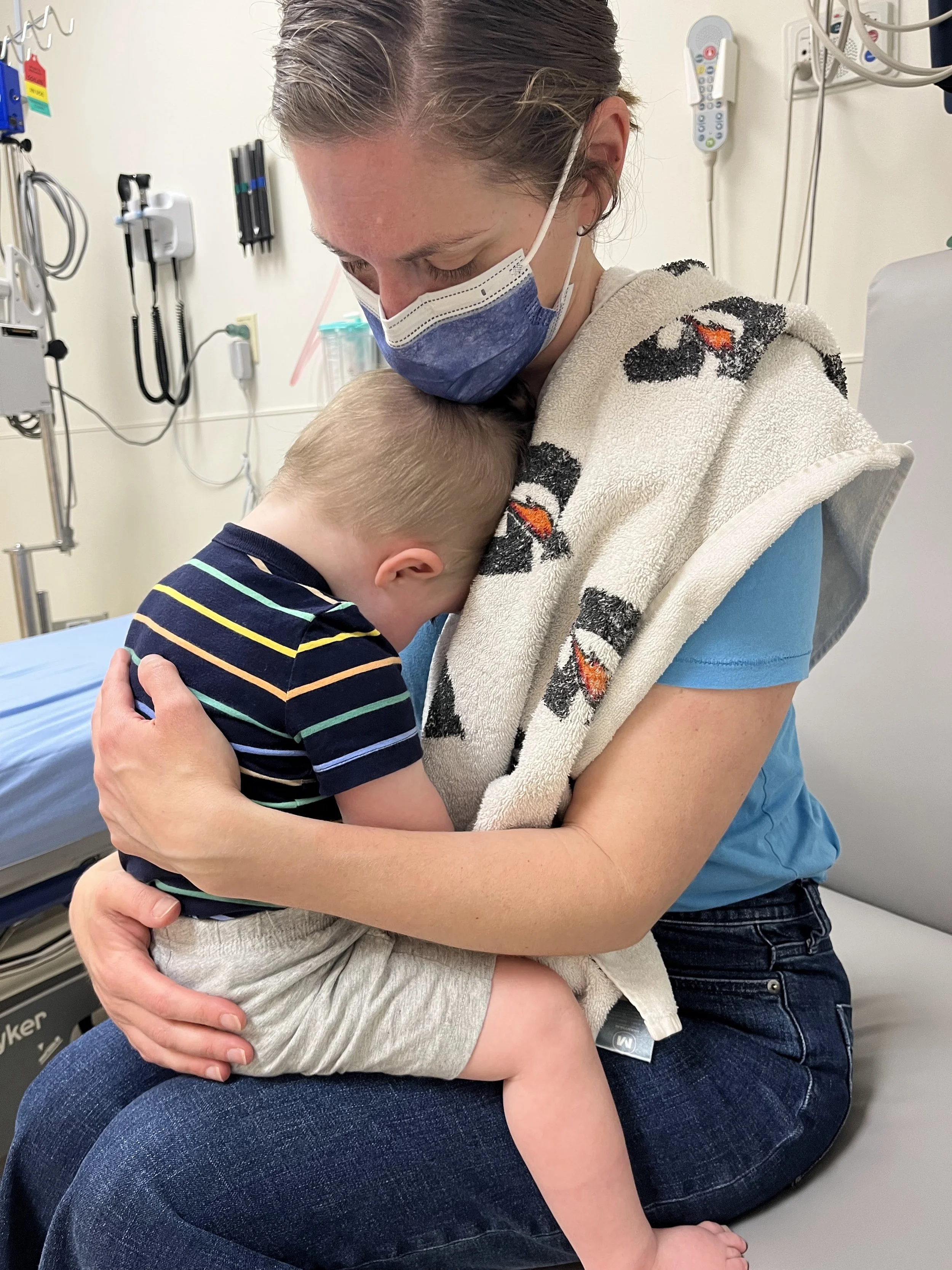 A woman wearing a face mask and a towel with penguin designs hugging a young boy in a hospital room.