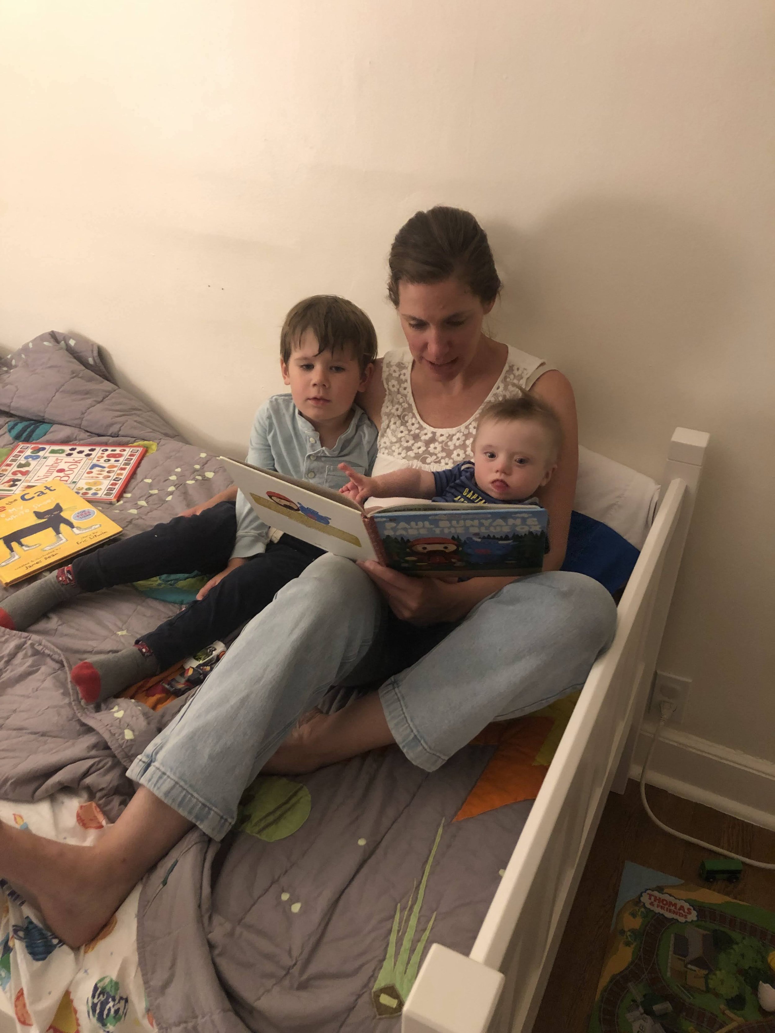 A woman reading a children's book to two young boys on a bed, with various children's books around them.
