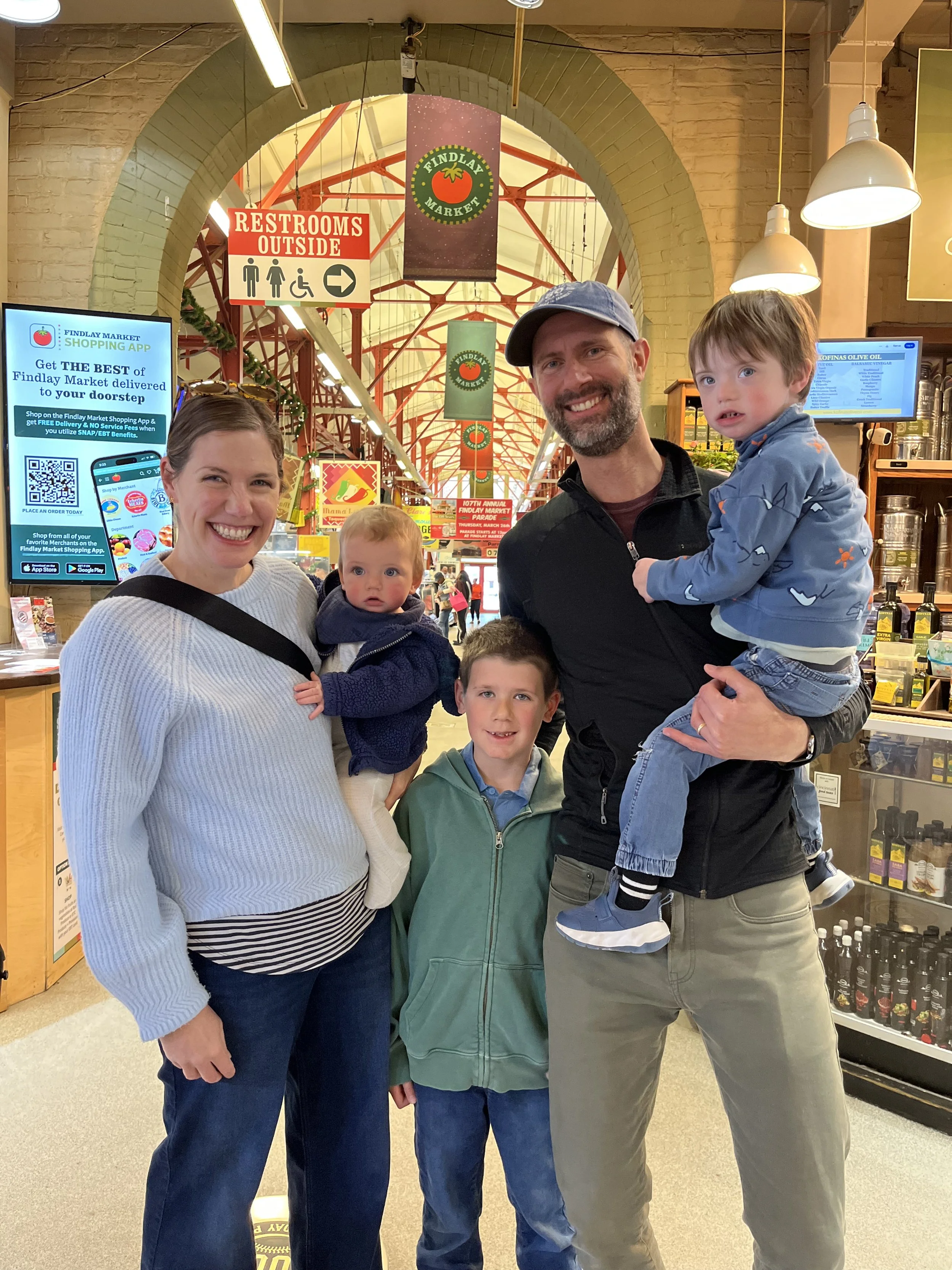 A family of four posing inside Findlay Market, with a woman, a man, and two young boys. They are smiling, and the man is holding one of the boys. There are signs for restrooms outside and a Findlay Market banner hanging from the ceiling.
