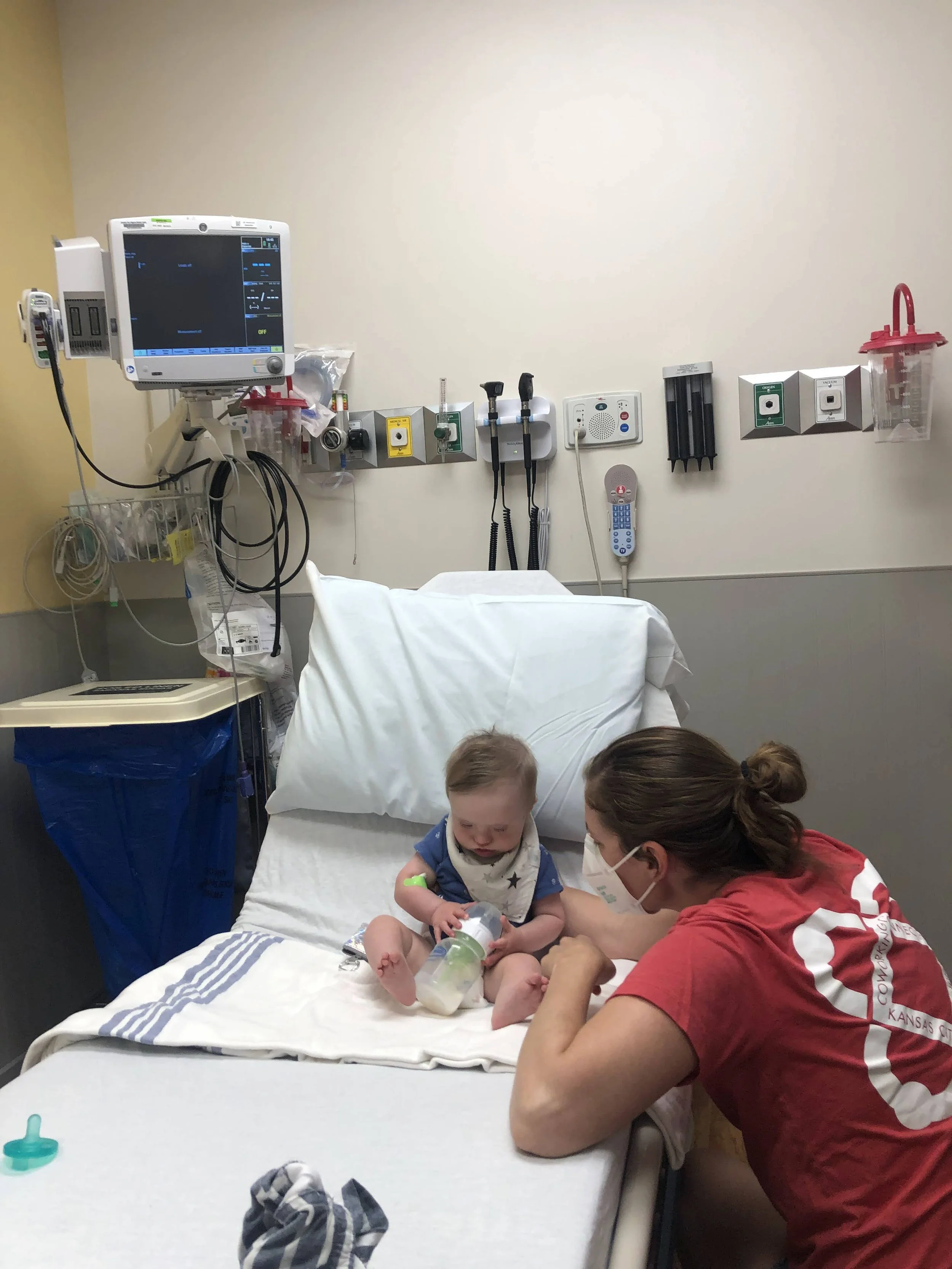 A young child with a feeding tube in a hospital bed interacting with a woman wearing a red shirt and mask, in a hospital room with medical equipment on the wall.
