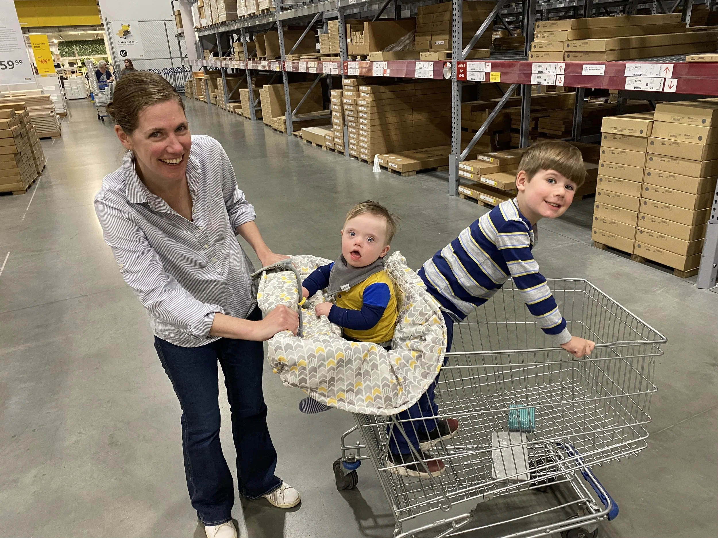 A woman with two young children in a shopping cart inside a home improvement store. The woman is smiling, pushing the cart, with a toddler sitting in a car seat in the front of the cart and an older child leaning over the side of the cart looking at 