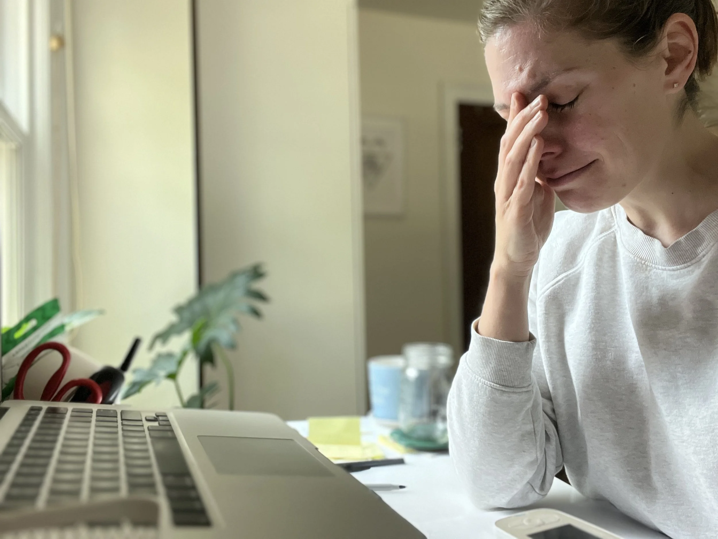A person with short gray hair and earrings, wearing a white shirt, sitting at a desk with a laptop, holding their forehead and looking down in a moment of frustration or fatigue.
