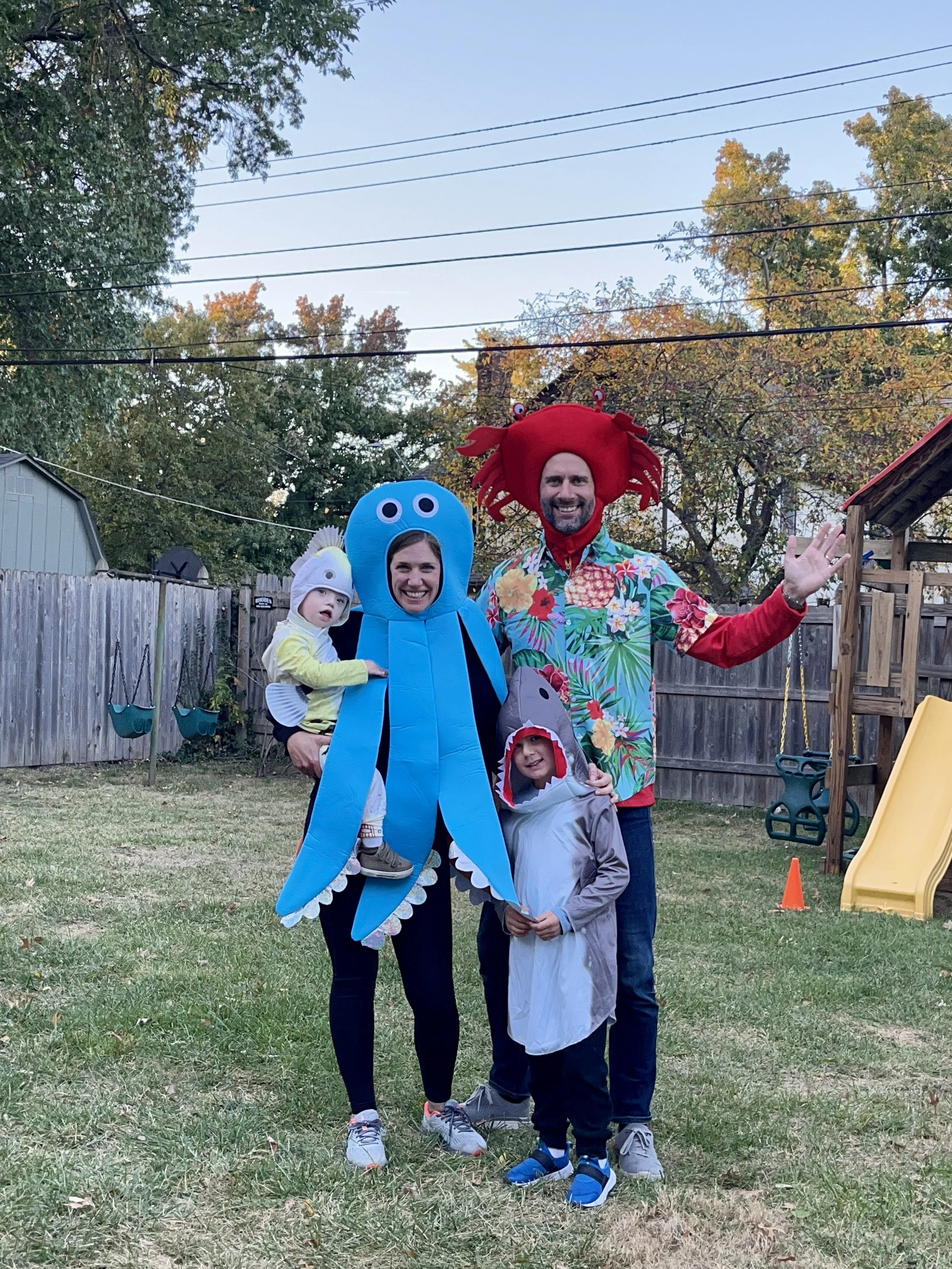 People dressed in crustacean, octopus, and shark costumes standing in a backyard with children, fence, trees, and a playground slide in the background.