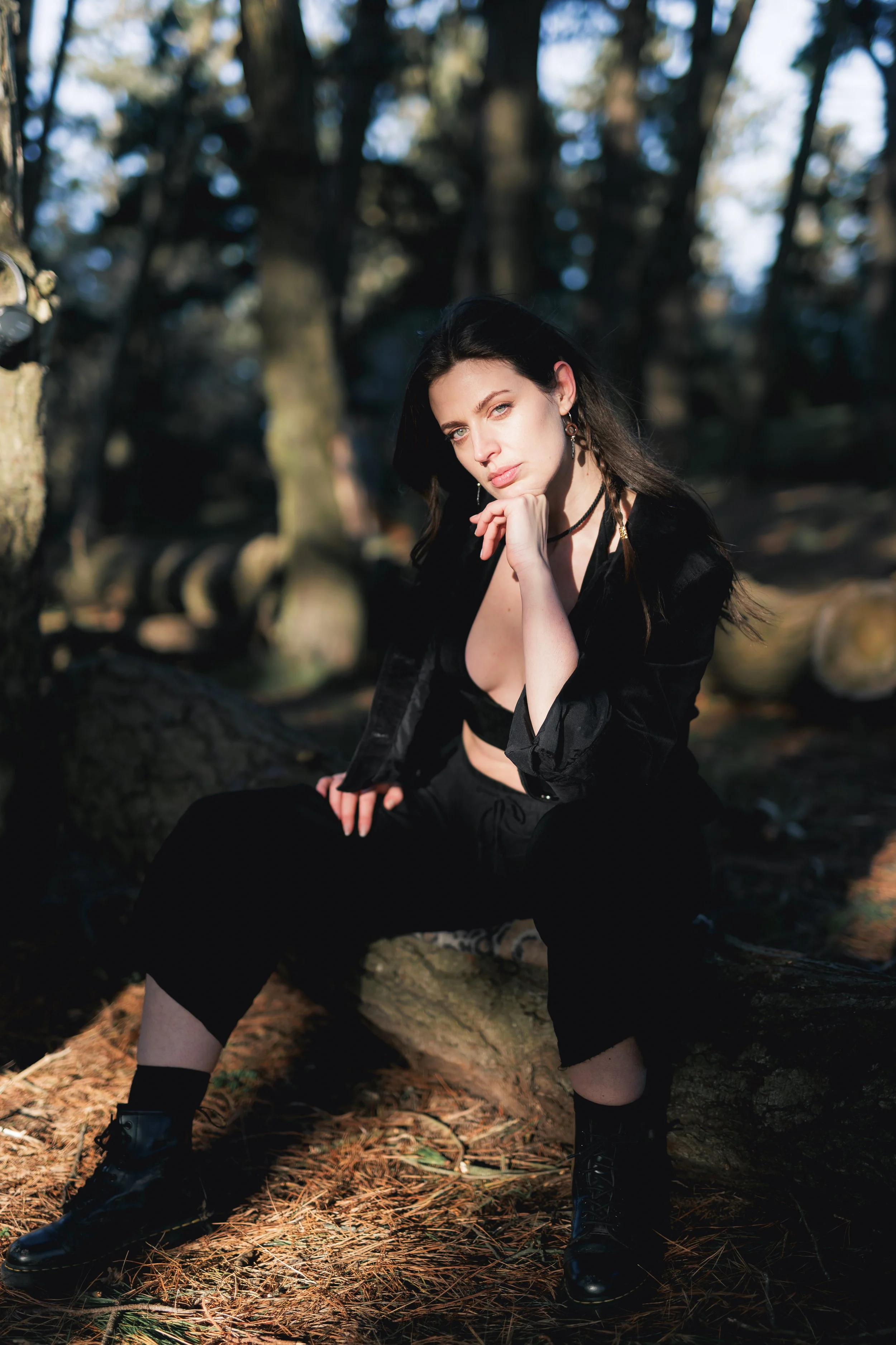 A woman with black hair and light skin sitting on a fallen tree trunk in a forest. She is wearing black clothing and boots, and has a thoughtful expression with her chin resting on her hand.
