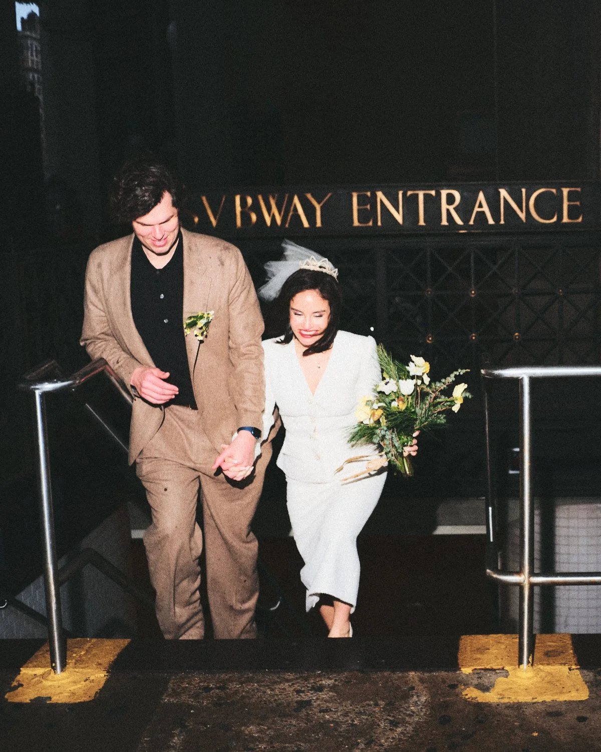 Documentary flash photography of a happy wedding couple holding hands. The woman is holding a bouquet of flowers, and both are smiling. Behind them is a sign for New York City Subway ENTRANCE and they are being photographed by Madivan Studios.