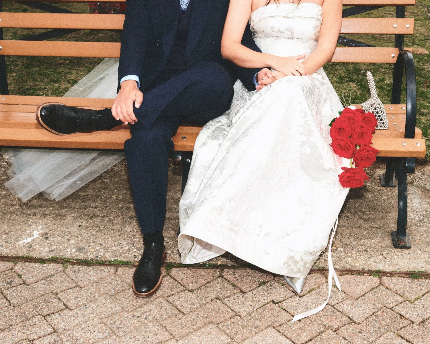 A couple sitting on a wooden NYC park bench, with the woman wearing a wedding dress and holding a bouquet of red roses, and the man in a navy blue suit, holding hands, photographed by Madivan Studios.