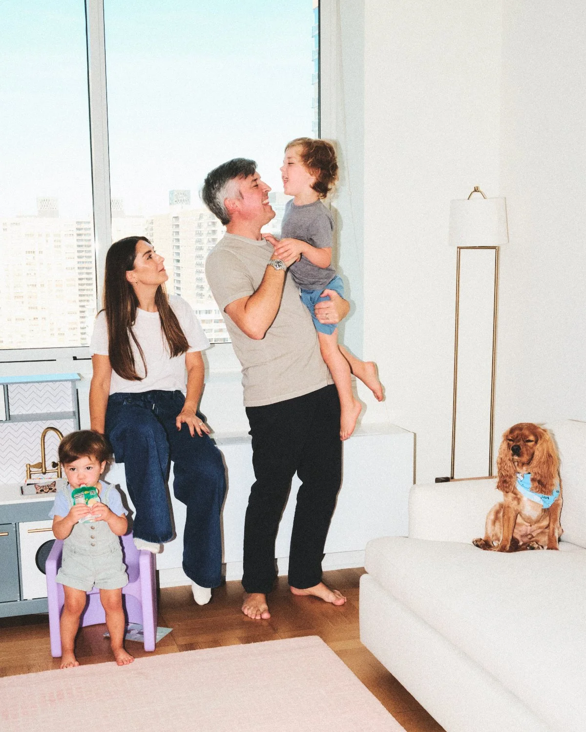 Documentary flash photography of a family with two children, one being held by the father, and a dog sitting on a couch in a bright living room with NYC skyline behind them and being photographed by Madivan Studios.