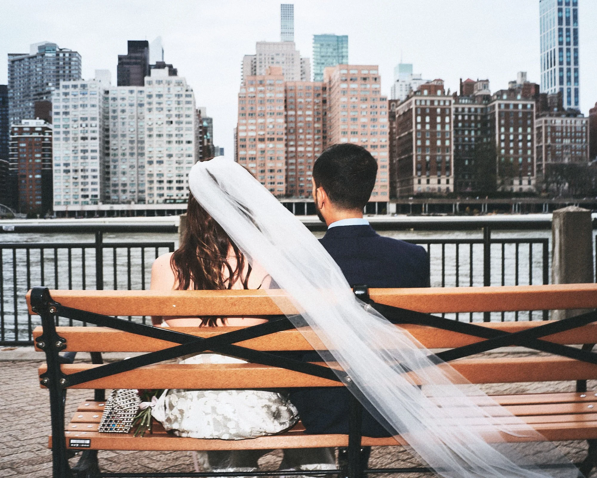 A bride and groom sitting on a park bench with the city skyline in the background, wedding bouquet and accessories placed on the bench.