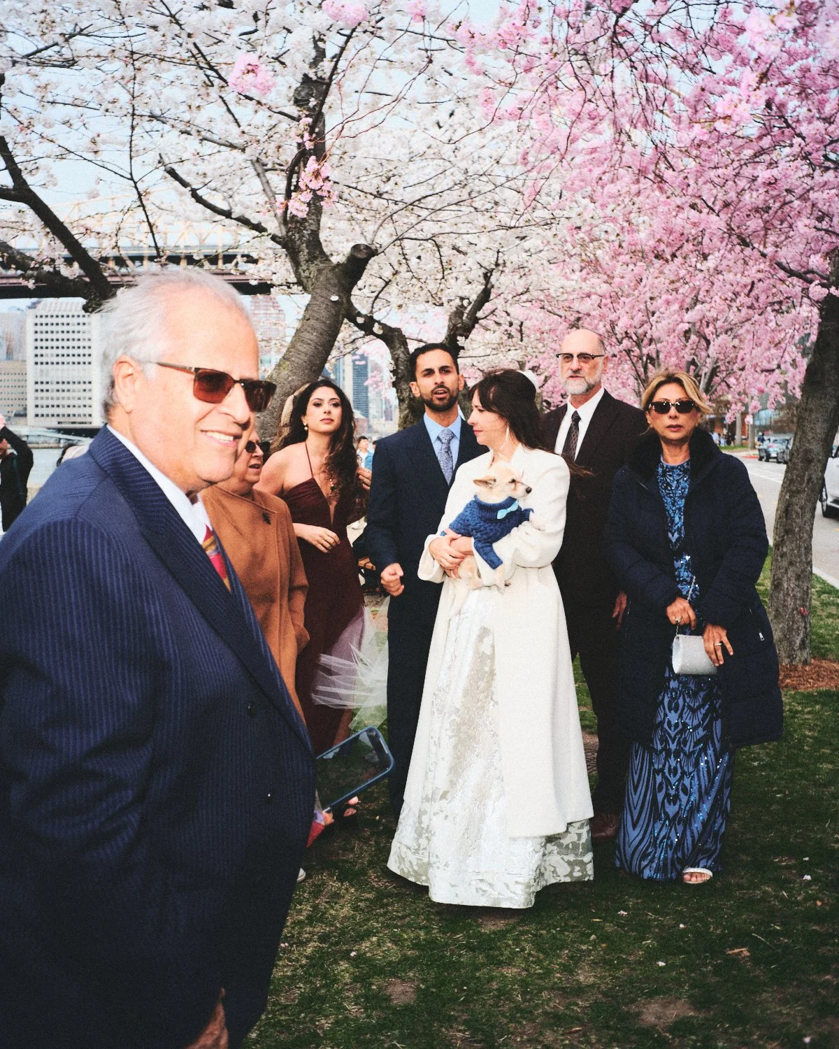 A group of people dressed formally stands under blooming cherry blossom trees outdoors with the New York City skyline in the background. Photographed by Madivan Studios.