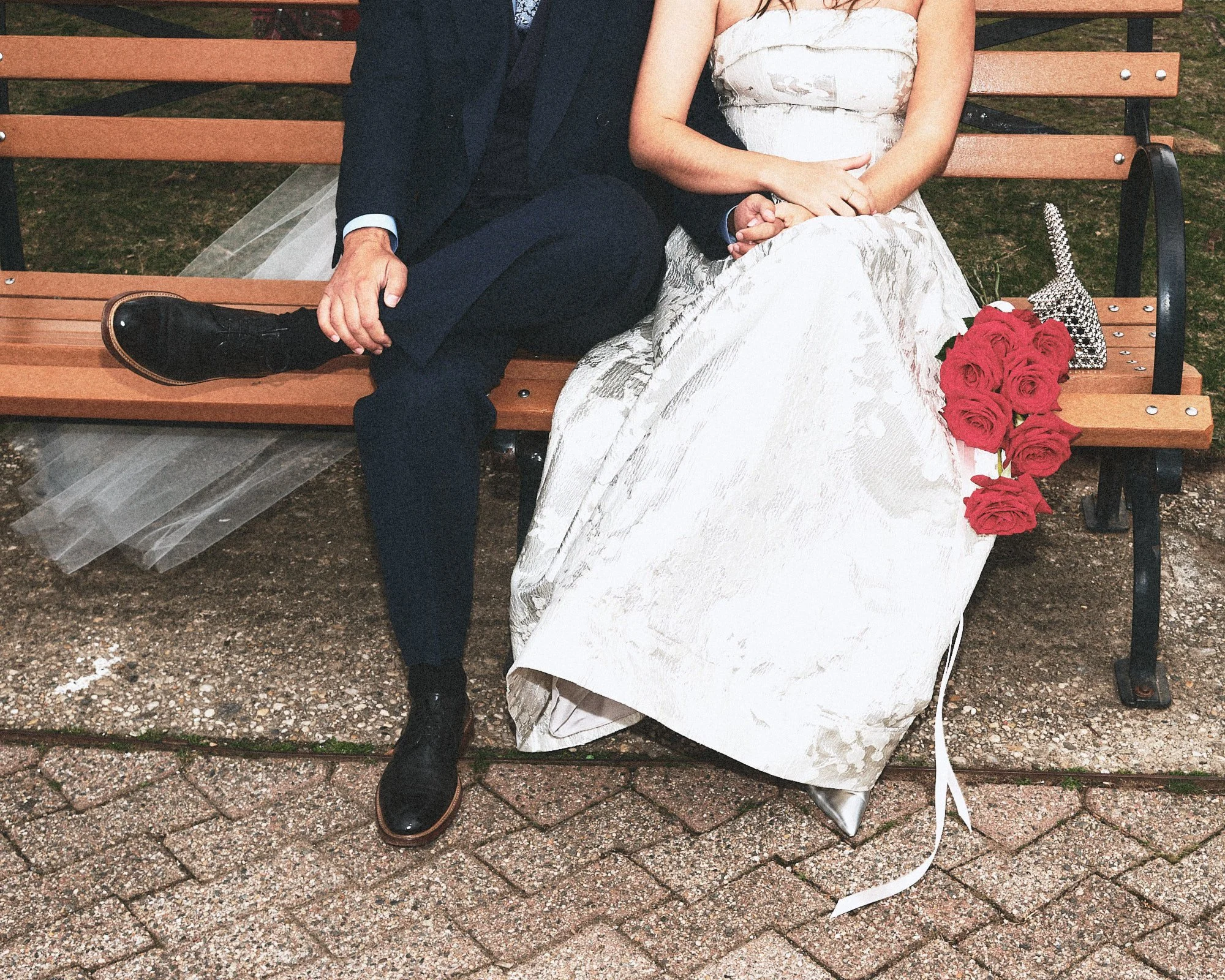 A man and woman sitting on a park bench, hand in hand, with a bouquet of red roses and a decorative silver elephant figurine placed beside them. The woman is wearing a white dress, and the man is dressed in dark clothing. The ground is covered with small pebbles.