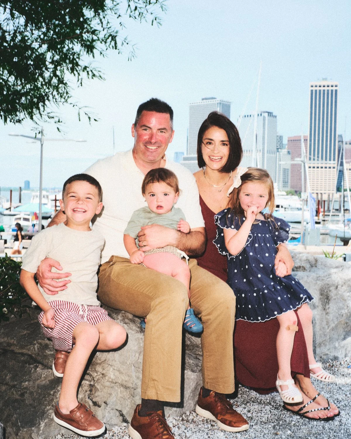 Documentary flash photography of a family of five sitting outdoors at the Dumbo marina with New York City skyline in the background, smiling and enjoying a sunny day and being photographed by Madivan Studios.