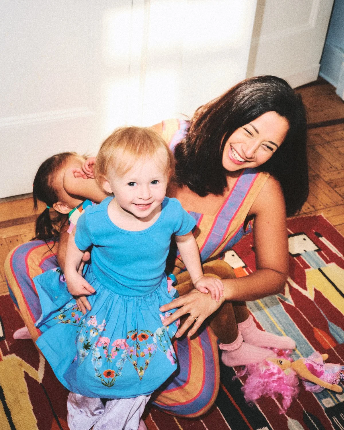 Documentary flash photography of a woman with dark hair and a colorful striped dress smiling while lying on the floor with two young children, a girl in a blue dress and short hair in NYC, near a patterned rug being photographed by Madivan Studios.