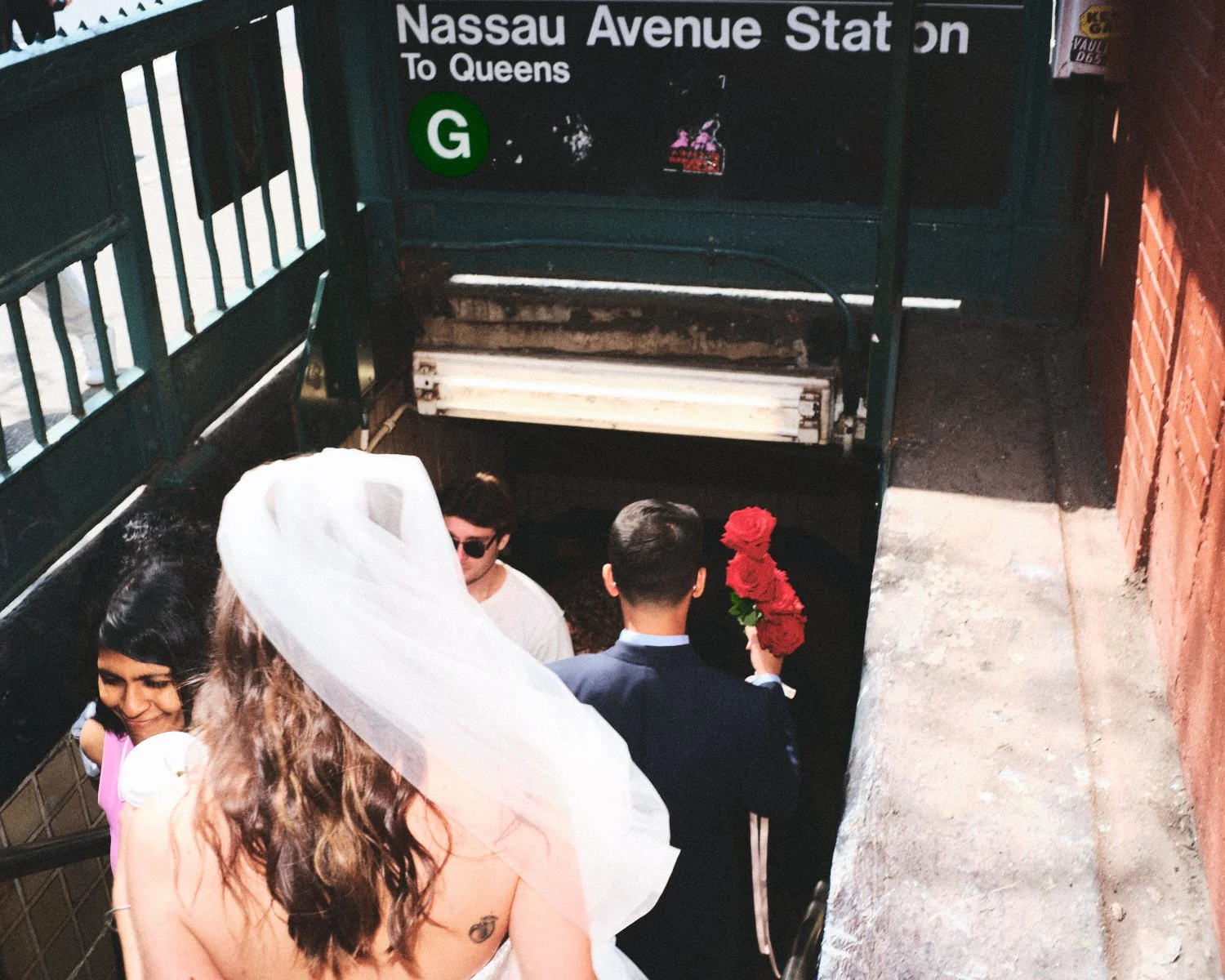 People descending into a subway station, with a sign indicating Nassau Avenue Station to Queens, including a woman holding red roses wearing a wedding veil, a man in a suit, and others, near stairs in an urban area photographed by Madivan Studios.