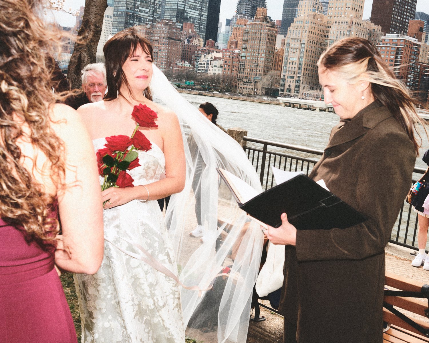 A wedding ceremony taking place outdoors near a river with the NYC skyline in the background. The bride is holding a bouquet of red roses, wearing a white dress photographed by Madivan Studios. A woman is reading from a book officiating the ceremony.
