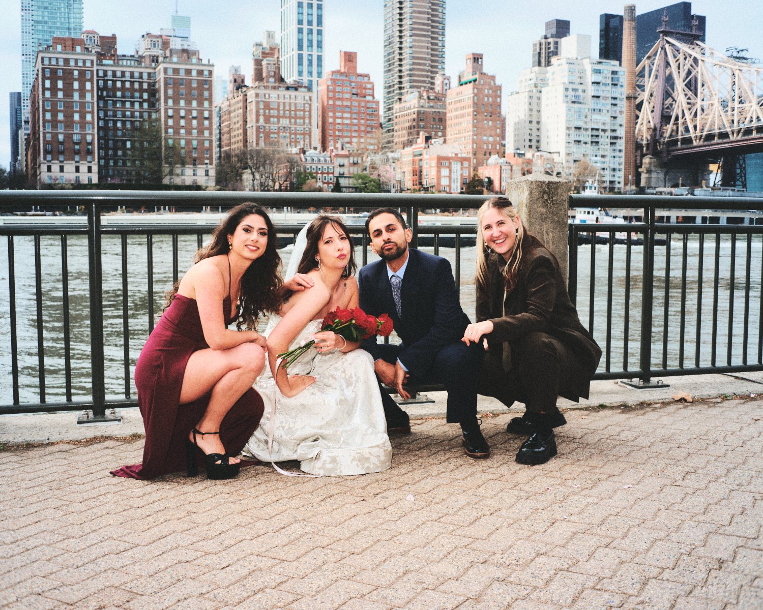 Four people photographed by Madivan Studios, including a bride holding a bouquet of red roses, posing by the water with the NYC skyline featuring tall buildings and a bridge in the background.