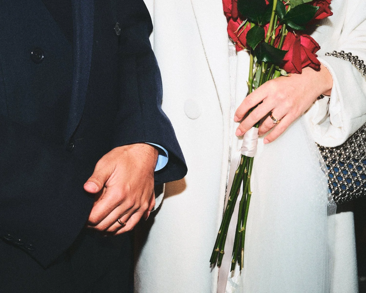Close-up of a couple's hands during a NYC wedding, with the bride holding a bouquet of red roses and the groom's hand resting on his lap. The bride is wearing a white dress with ornate embellishments, and the groom is dressed in a dark suit.