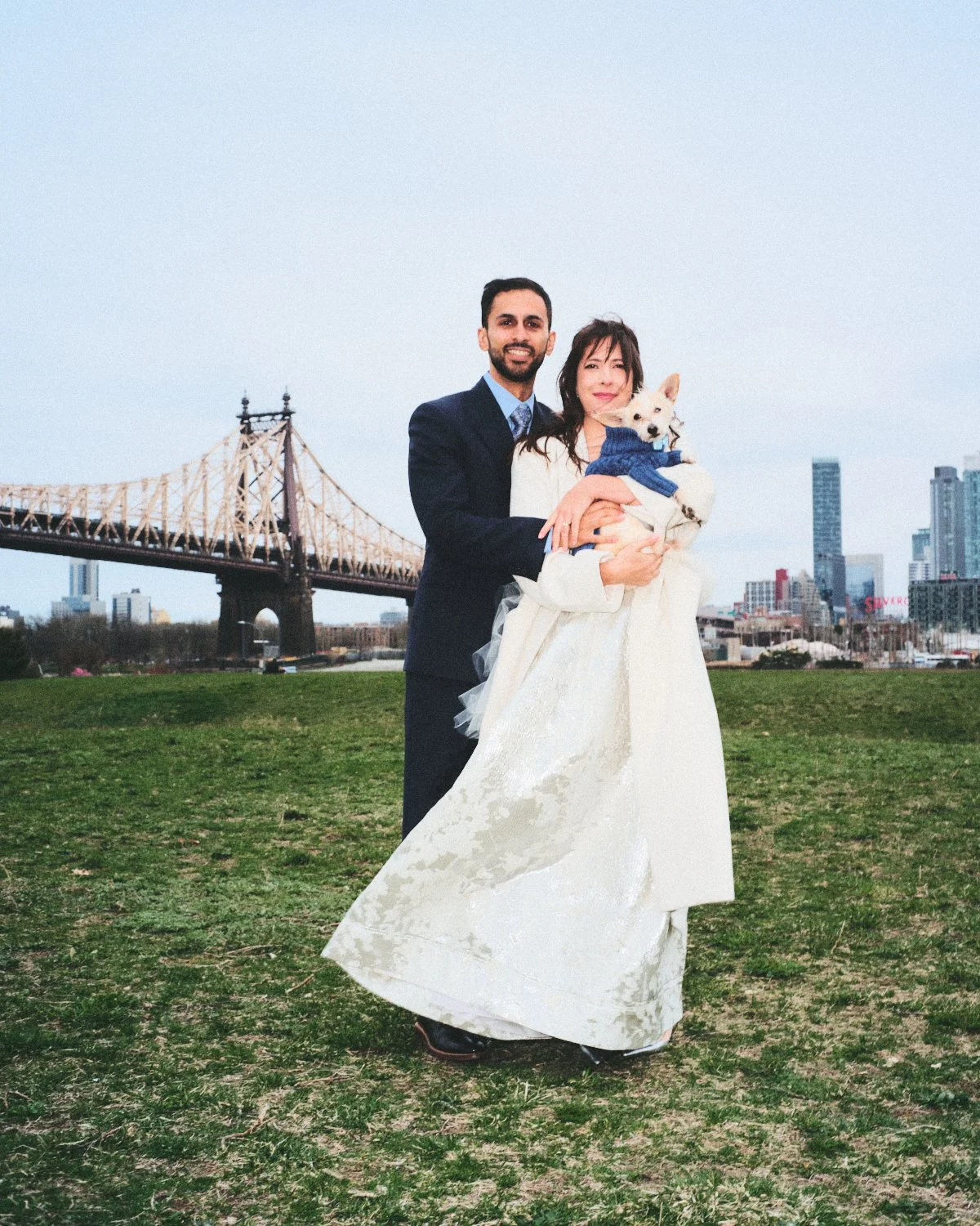 A smiling couple dressed in wedding attire standing outdoors on a grassy area, holding a small white dog with a blue sweater, in front of a bridge and New York City skyline, photographed by Madivan Studios.