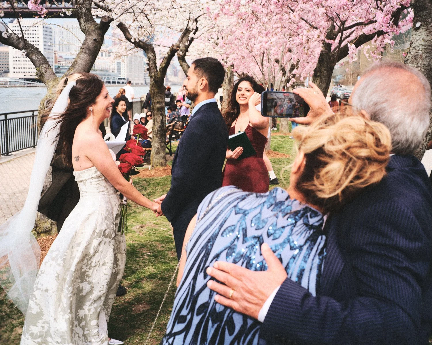 A wedding ceremony taking place outdoors near pink cherry blossom trees with NYC cityscape in the background photographed by Madivan Studios. The bride and groom are facing each other, holding hands, while guests look on and take photos.