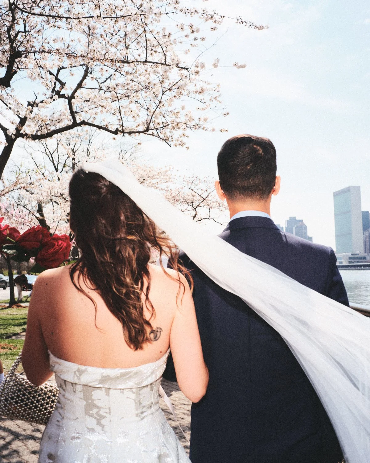 A couple dressed in wedding attire photographed by Madivan Studios, with the woman wearing a white dress and the man in a dark suit, standing outdoors near cherry blossom trees and the NYC skyline.
