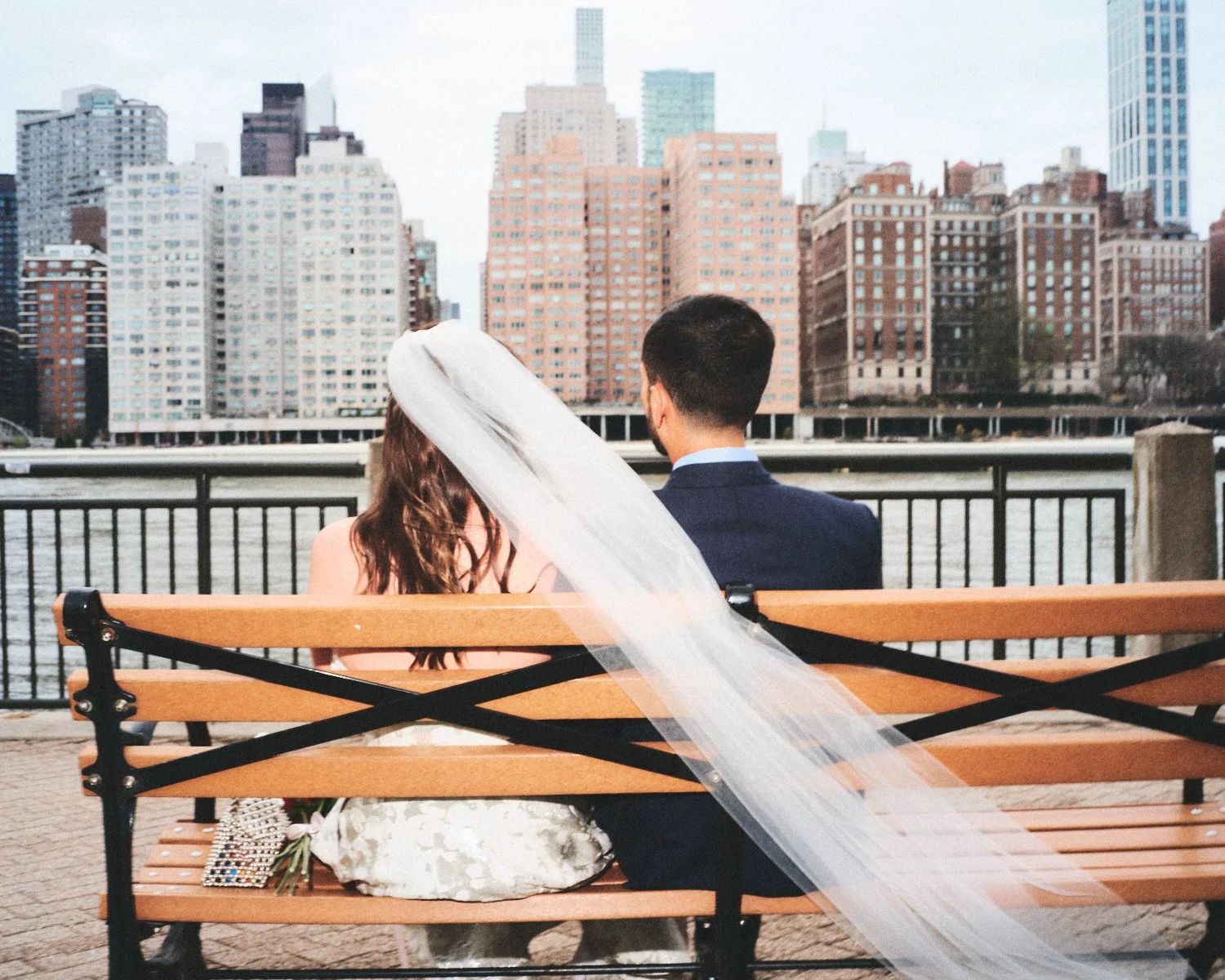 A bride and groom sitting on a NYC park bench facing a river and city skyline, with the bride wearing a wedding dress and veil, and the groom in a dark suit. Photographed by Madivan Studios.