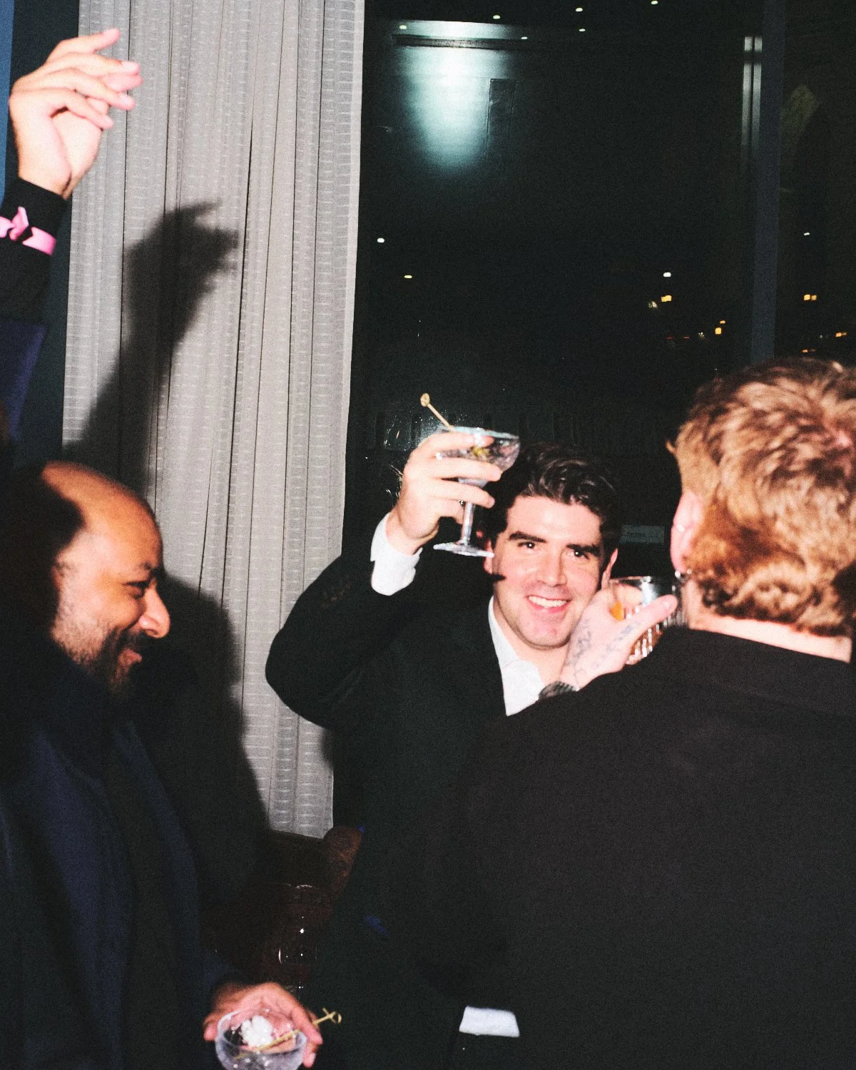 Documentary flash photography of a group of men at a corporate event celebration, with one man in the center raising a cocktail glass and smiling, they are being photographed by Madivan Studios..