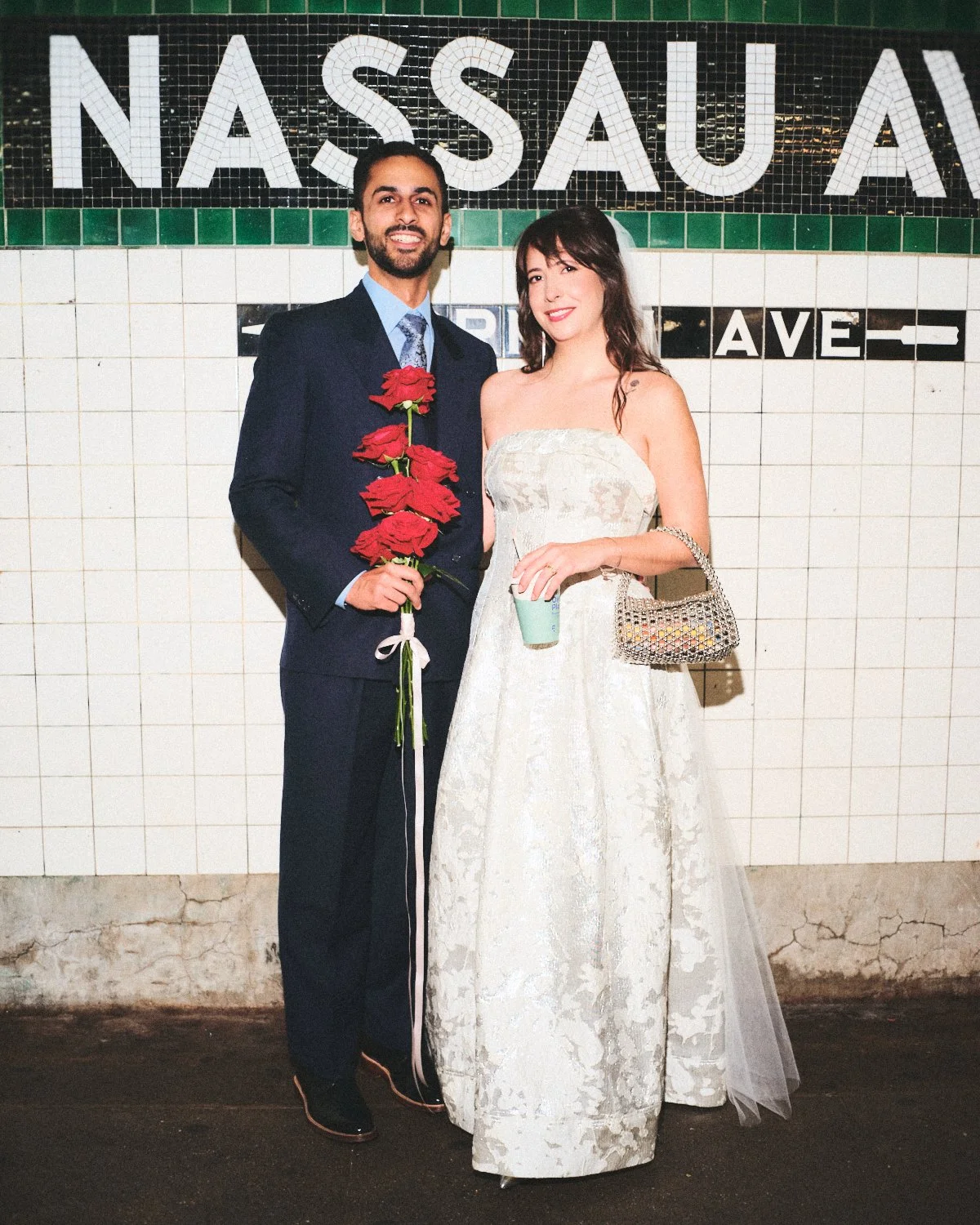 A man and woman in formal attire standing in front of a NYC subway station sign photographed by Madivan Studios. The man is holding a bouquet of red roses and the woman is holding a drink and a small handbag.