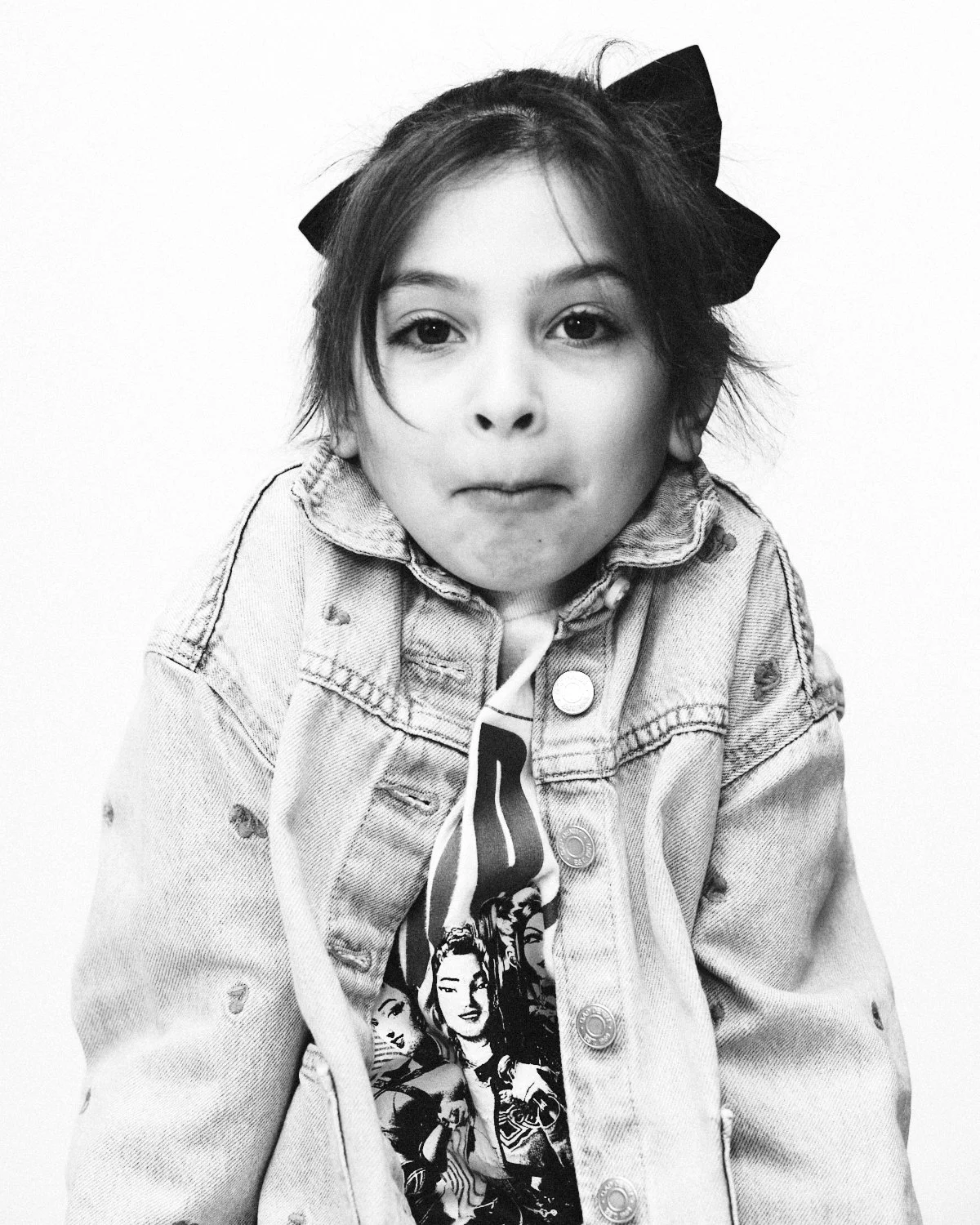 Black and white photo of a young girl wearing a denim jacket with a comic book or graphic T-shirt underneath and a large bow in her hair. Photographed in monochrome by Madivan Studios in New York City.