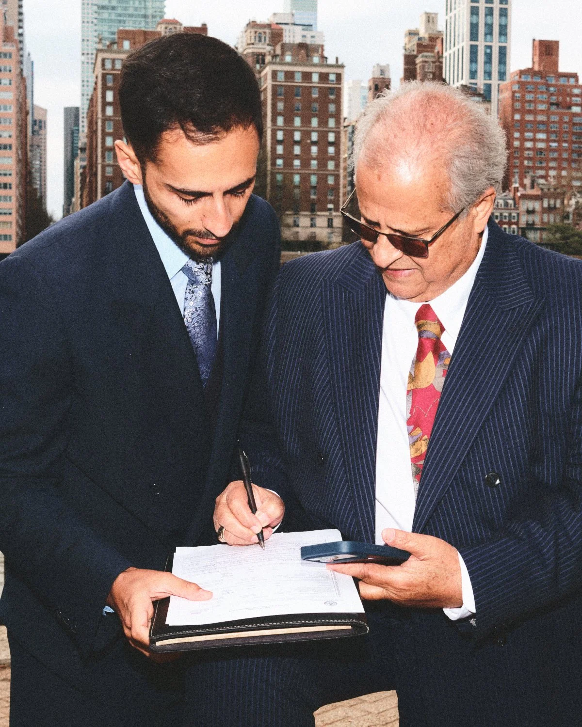 Two men in business suits reviewing documents and a mobile phone against the New York City skyline with tall buildings photographed by Madivan Studios.