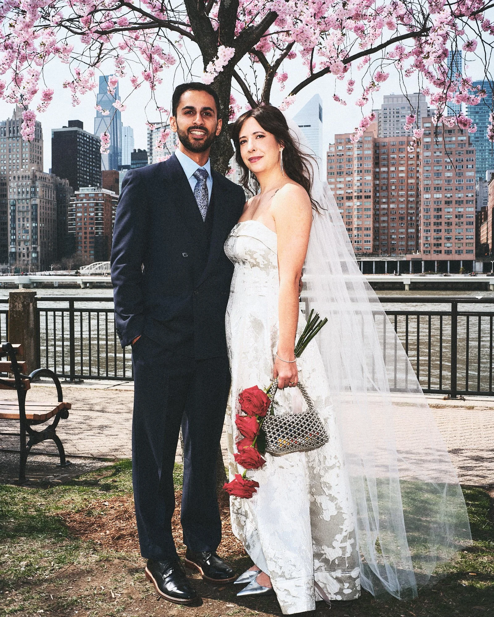 A newlywed couple standing under a pink blossoming tree with city buildings in the background. The groom is dressed in a dark suit with a light blue shirt and patterned tie, and the bride is in a white lace strapless wedding gown holding a bouquet of red roses and a small black and white purse, with a sheer veil.