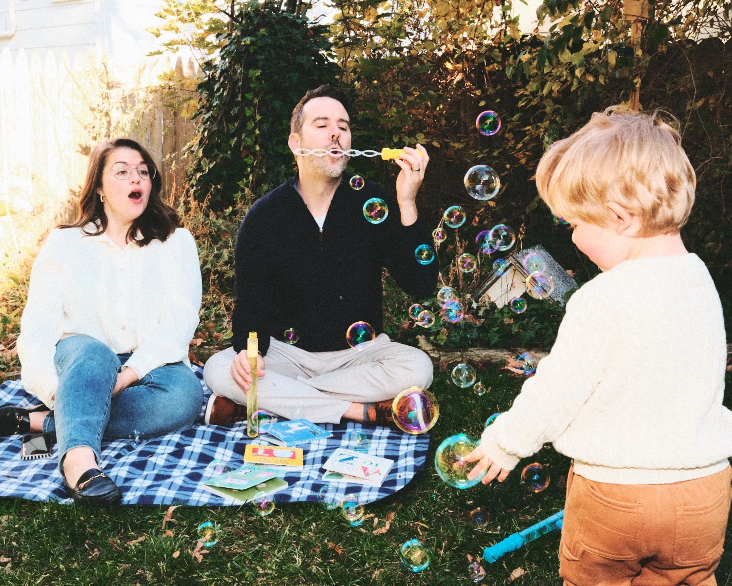 Documentary flash photography of two adults and one child, sitting on a blue checkered blanket outdoors with a garden background in New York City. The man is blowing bubbles while the woman and child are being photographed by Madivan Studios.
