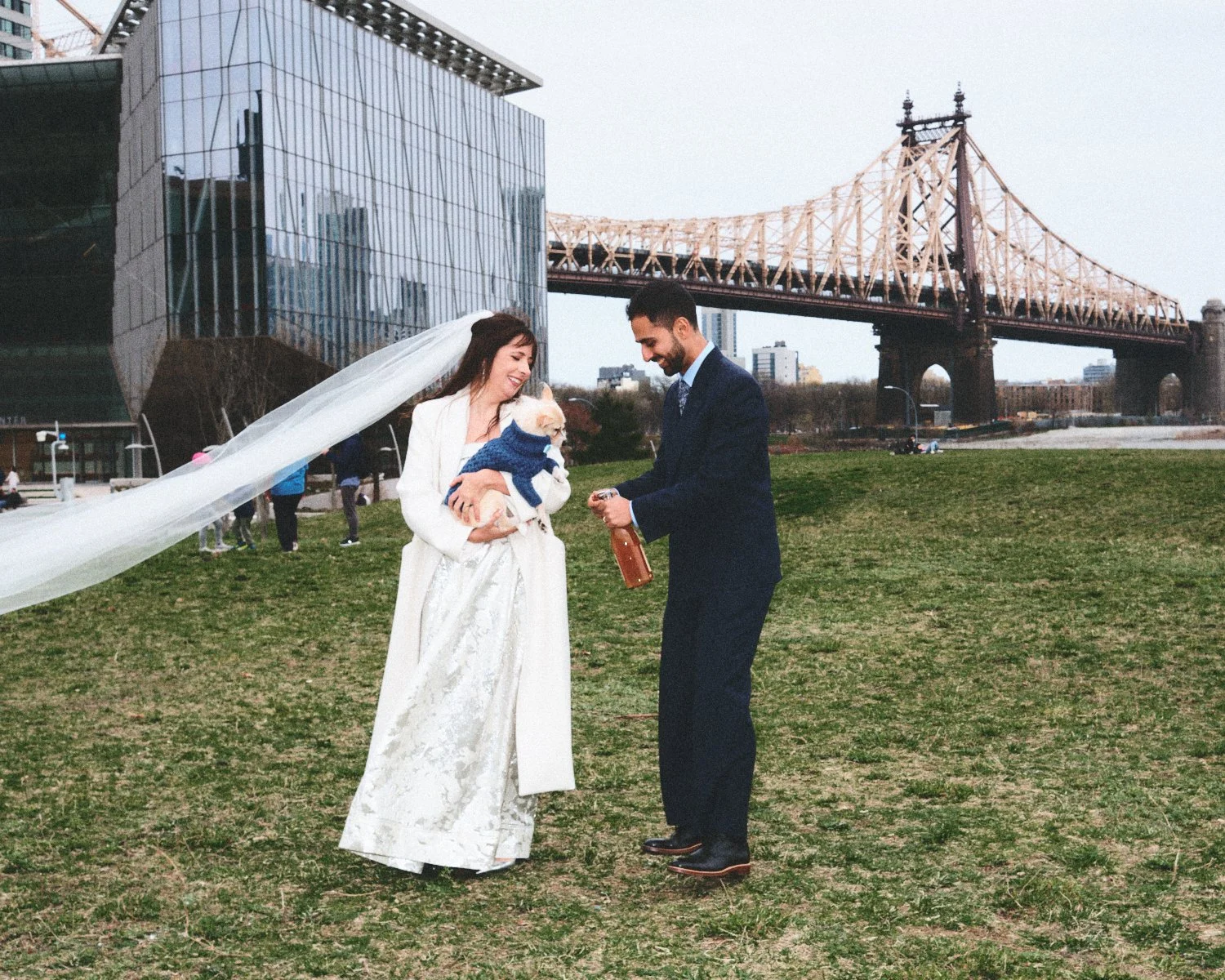 A wedding scene in an outdoor park with a bride in a white dress holding a small dog in a blue sweater, standing with a groom in a dark suit. A veil is flowing from the bride's head. NYC bridge and photographed by Madivan Studios.
