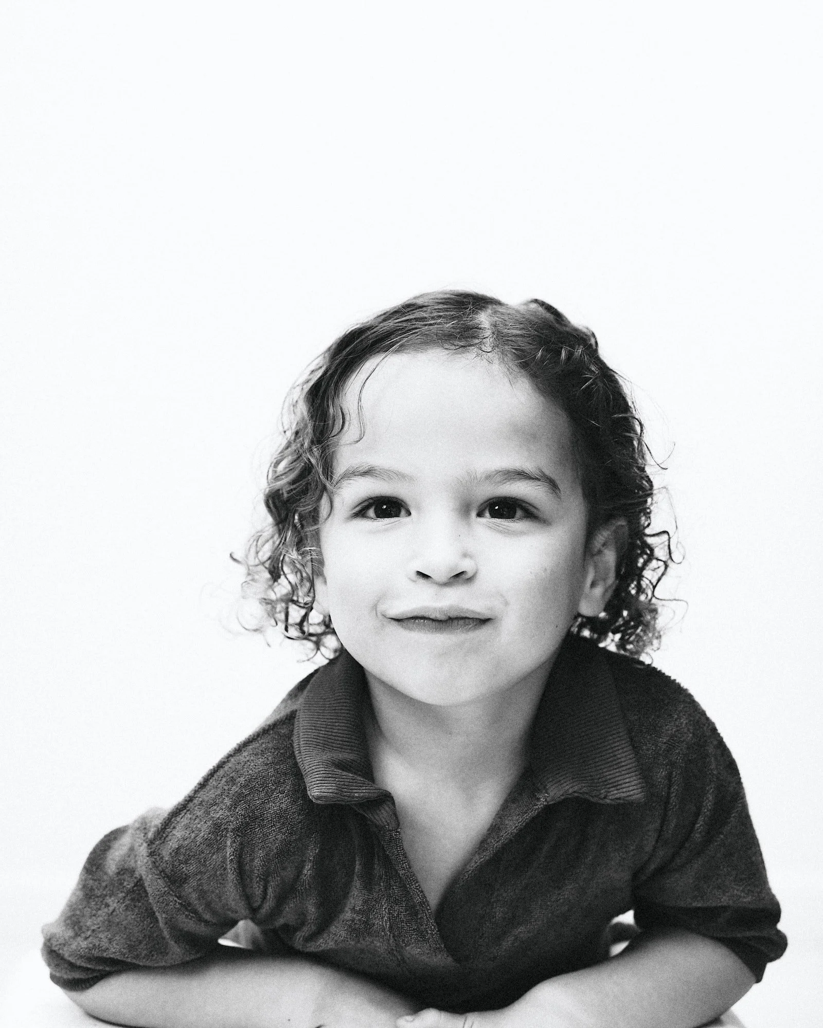 Black-and-white photo of a young girl with curly hair, smiling, wearing a collared shirt and sweater, leaning on her forearms.
