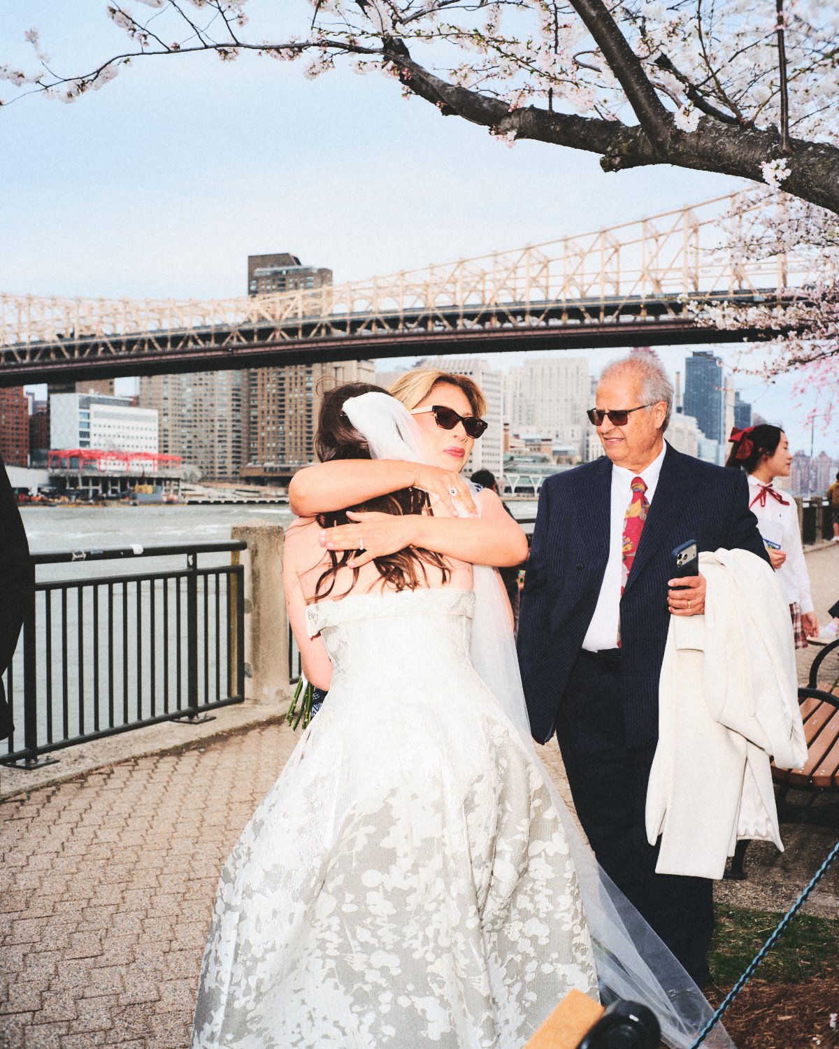 A woman in a wedding dress and sunglasses hugging another woman, while a man in a suit and sunglasses looks on holding a coat photographed by Madivan Studios. They are outside with the NYC skyline, bridge, and cherry blossom trees in the background.